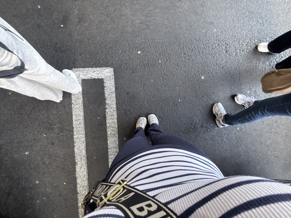 The scene depicts an overhead view of four individuals standing on dark grey asphalt pavement featuring a white painted rectangular ground marking. The precise location within Culver City, United States of America, is not identifiable from the visual content.
One individual, positioned on the lower left, wears a white and navy blue striped garment, navy blue pants, and light-colored athletic shoes. A black and white strap with gold-colored hardware and partial "BI" lettering is visible across their torso. Their feet are positioned adjacent to the left side of the white rectangular marking.
To the top right, a second individual's lower body is visible, wearing light grey sweatpants and white athletic shoes with a visible black Nike swoosh. Their right foot is placed inside the white rectangular line. A black strap is visible over their shoulder.
A third individual is located in the bottom-right quadrant, wearing blue jeans and white athletic shoes. A tan handbag rests on the ground near their feet.
The lower legs and white athletic shoes of a fourth individual, wearing dark pants, are partially visible at the far bottom-right.
All subjects are standing. No explicit actions or interactions beyond their shared presence on the pavement are depicted. The asphalt surface shows minor texture and scattered light-colored debris.