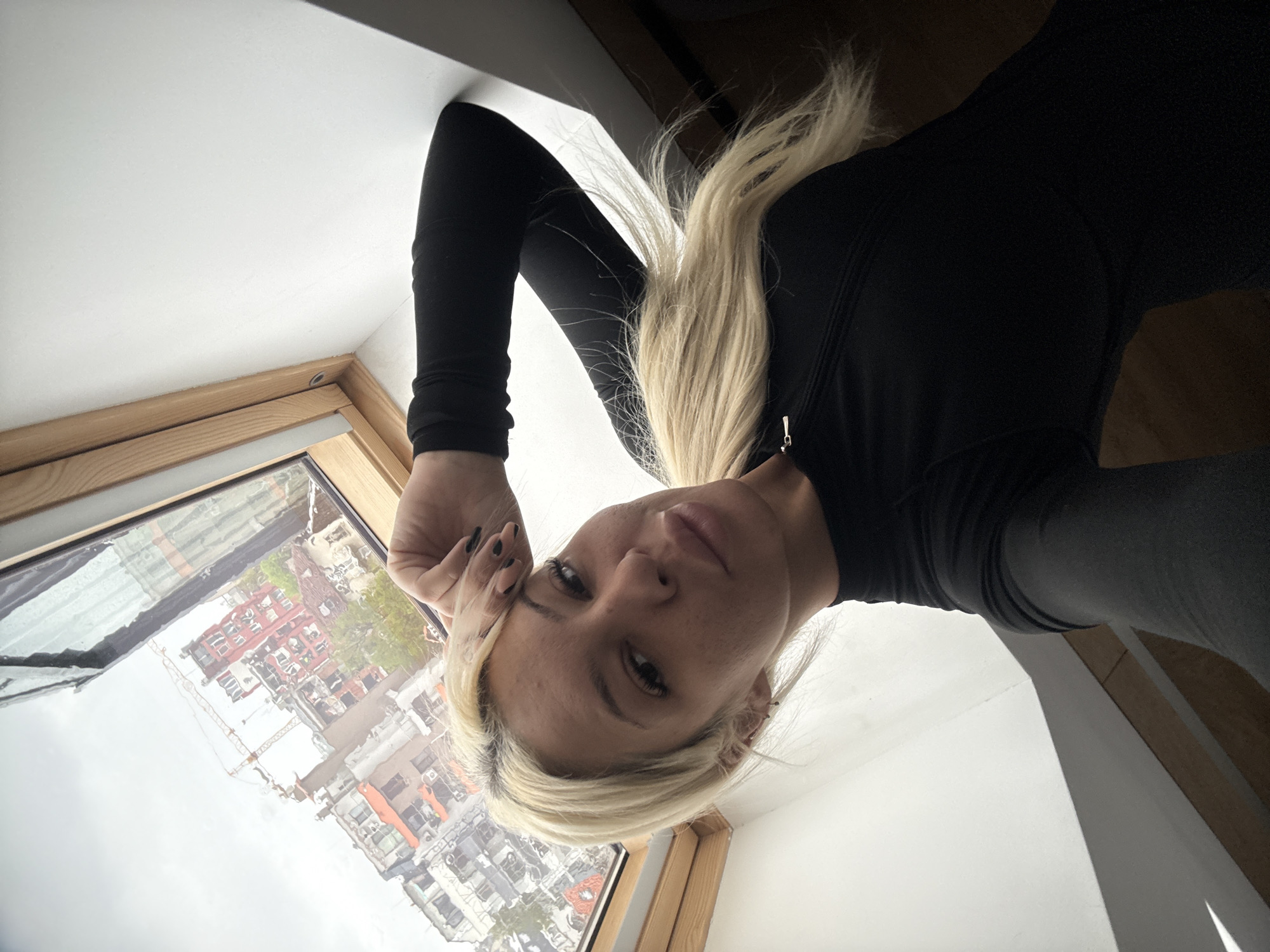 A woman with long blonde hair, wearing a black long-sleeved top, takes a selfie. Her body is positioned upside down in the frame, suggesting she is leaning over, looking downwards or outwards. She has a neutral expression and looks directly at the camera. Her hand, with dark nail polish, rests near her temple.
The scene is indoors, likely a room with a sloped ceiling or an attic, featuring a wooden-framed window, possibly a skylight or roof window. The window reflects an urban environment, showing numerous multi-story buildings with varied architecture, including some with red roofs and others with light-colored facades. A tall construction crane is also visible in the reflection, indicating a bustling city. The lighting suggests it is daytime, possibly overcast. Parts of a white wall or ceiling and a dark wooden surface are visible around the woman.
The location is Sofia, Bulgaria. No text is visible in the image.