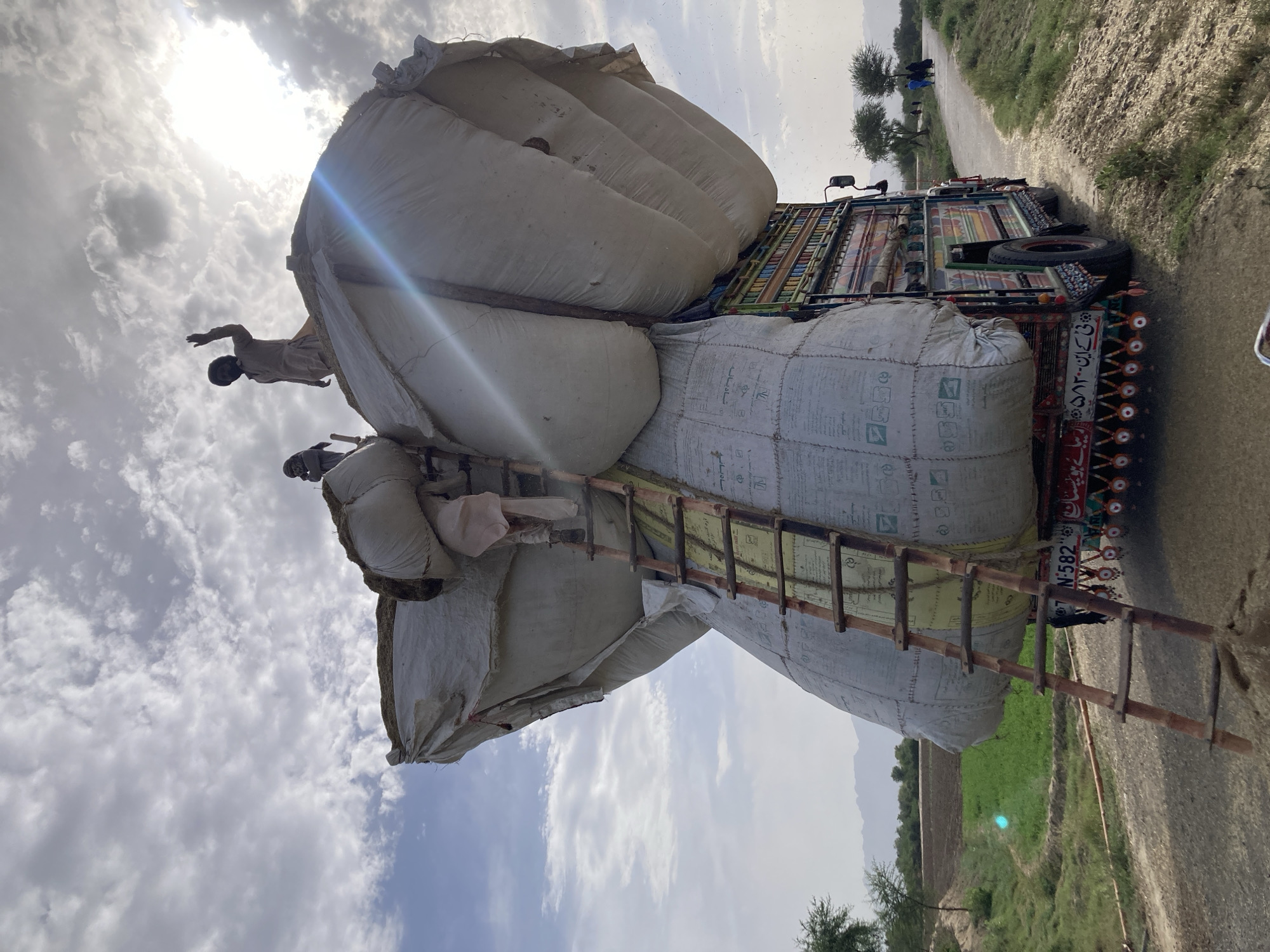 Two men are on top of several large sacks on a truck in Rāwāni, Pakistan. A wooden ladder leans against the sacks. The truck is decorated with colorful designs. The background includes a road, fields, and a cloudy sky with the sun shining through.
