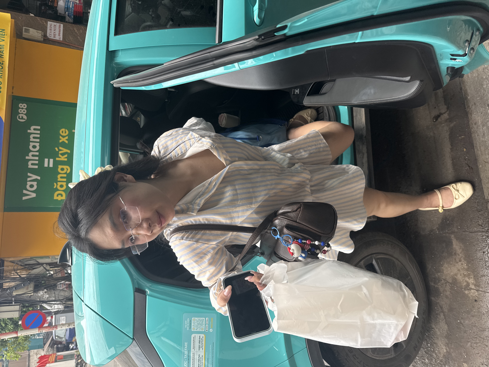 A young Vietnamese woman with glasses and her hair pulled back is stepping out of a turquoise car with the door open. She is wearing a light-colored pajama-style outfit with blue and yellow stripes. In her right hand, she holds a white plastic bag and a black smartphone. She has a small brown purse slung over her shoulder, adorned with a keychain featuring a character that resembles Hello Kitty. Her left leg is bent, with her foot resting on the ground. She is wearing beige flat shoes with bows. The car appears to be a taxi, as indicated by a sticker on the rear passenger door with Vietnamese text that includes "TAXI FARE". The scene is set on a street in Hanoi, Vietnam, with a yellow and green sign in the background featuring Vietnamese text and a logo that says "F88", likely indicating a financial institution or service. The ground is wet and reflective, suggesting recent rain.