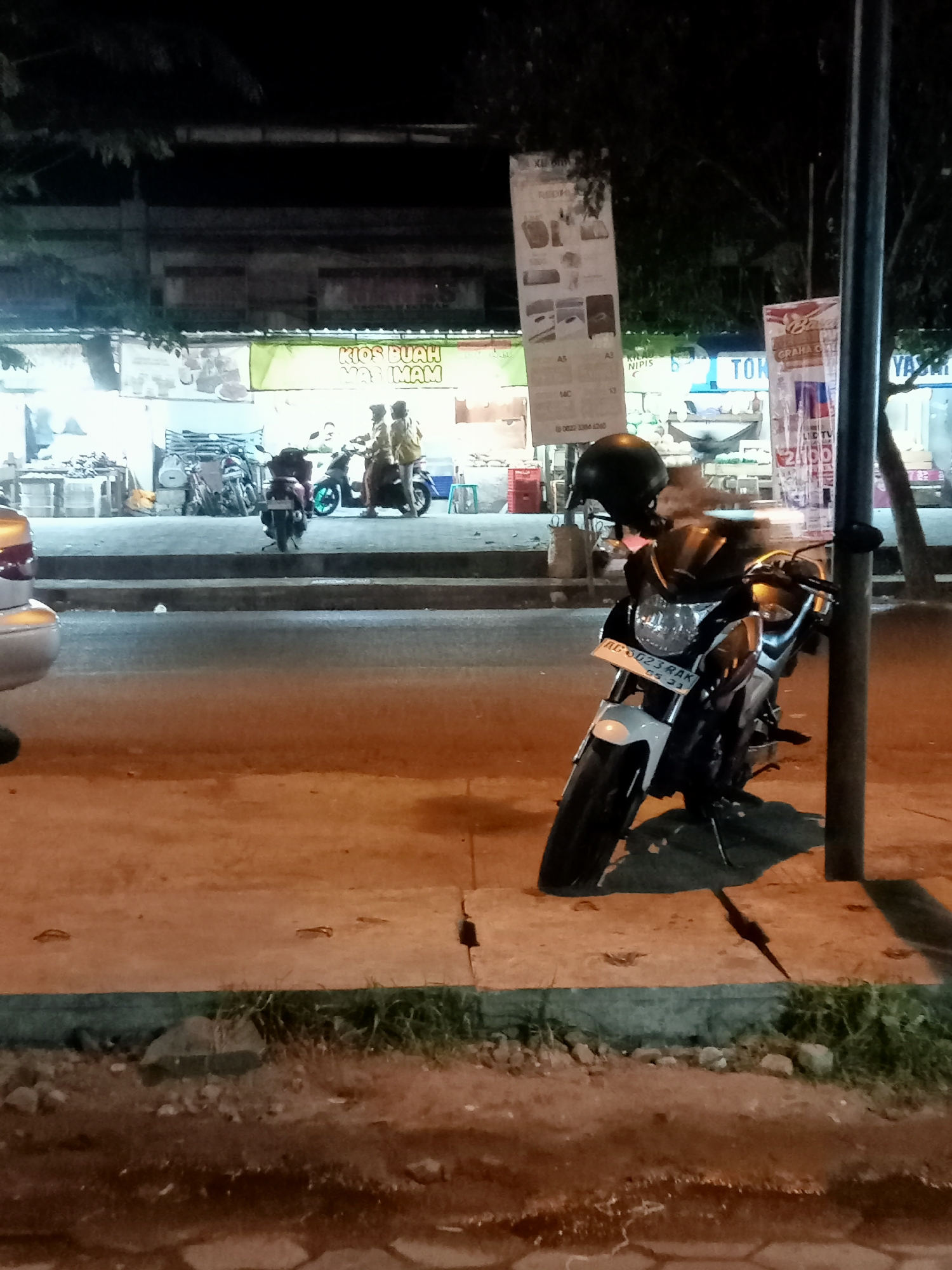 A nighttime scene is depicted on a street in Mangunsari, Indonesia, with a focus on a parked motorcycle. The motorcycle, white and black, is in the foreground, leaning on its kickstand. It has a license plate reading "AG 5023 RAK". A black helmet rests on its handlebars. In the background, a row of buildings with signs is visible, suggesting commercial establishments. One sign, in green and yellow, reads "KIOS BUAH MAS IMAM". Several other motorcycles and individuals are present further down the street, some appearing to be standing or on their bikes. The lighting is artificial, casting shadows and highlighting the urban environment. The ground consists of pavement and a dirt path with grass and rocks.