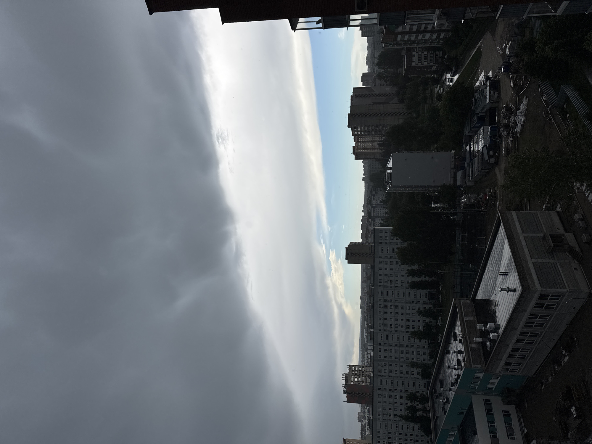 The image captures a high-angle, slightly tilted view of an urban landscape. A dense residential area, characterized by high-rise buildings with repetitive window patterns, stretches towards the horizon. On the right side of the frame, what appears to be a construction or storage area is visible, filled with containers and other materials. A stark contrast is presented by the heavy, dark, overcast sky on the left, which dominates a significant portion of the image. A narrow strip of blue sky can be seen on the middle, between the dark clouds and buildings. The buildings and ground below appear damp, suggesting recent rainfall. The overall atmosphere is gloomy and overcast.