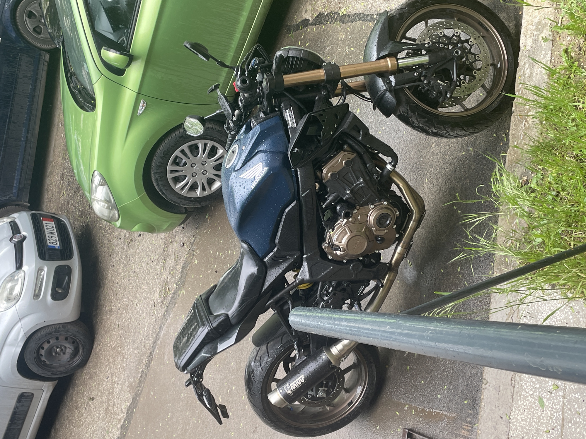 The image shows a blue Honda motorcycle parked on an urban street next to a green car and a white Fiat Panda, in what appears to be an outdoor setting. The motorcycle is wet, suggesting recent rain. The background suggests a cityscape. The license plate on the Fiat suggests the location of Sofia, Bulgaria.