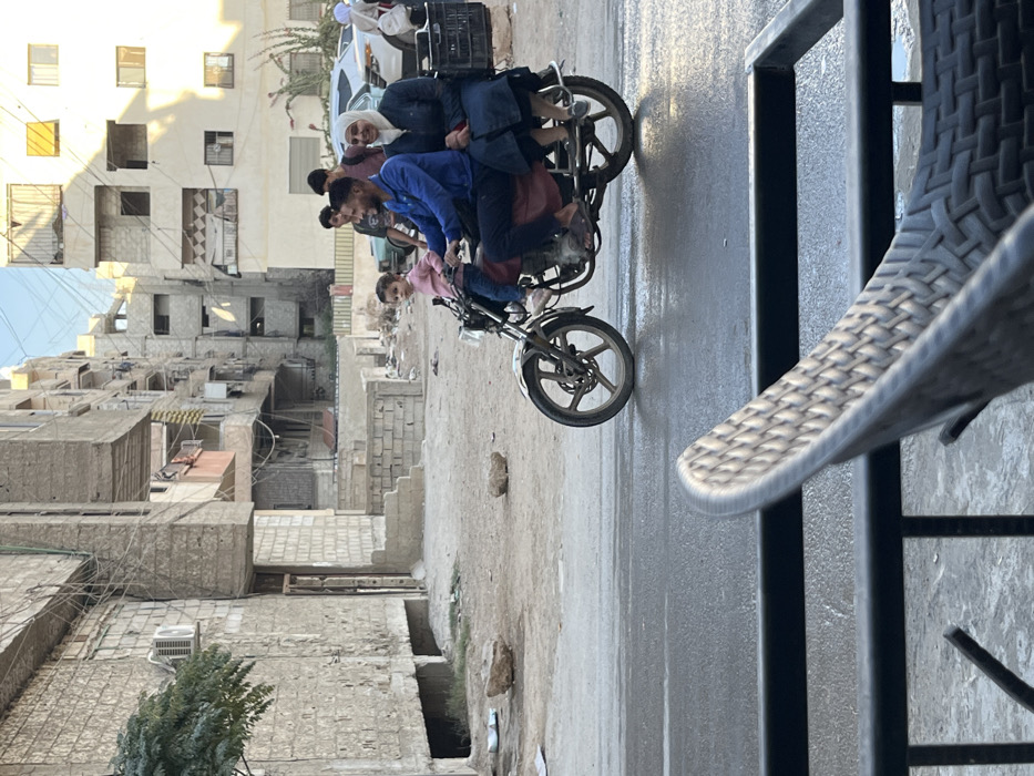 The image depicts an urban street scene in Mu'adammiyat ash-Sham, Syria, captured from a slightly elevated perspective.
In the midground, a family of three rides a dark-colored motorcycle from left to right on a wet or reflective asphalt road. The driver is an adult male wearing a blue long-sleeved shirt, dark pants, and sandals, smiling and looking slightly away from the camera. A young child, dressed in a pink long-sleeved top and dark pants, sits directly in front of the driver, facing forward with a neutral expression, looking towards the camera. An adult female, wearing a white hijab and a dark blue dress, sits behind the driver, also in sandals, smiling and looking slightly away from the camera. A black plastic crate is attached to the rear of the motorcycle.
The background features a dirt and gravel shoulder adjacent to the road, littered with a few rocks. Beyond this, several multi-story residential buildings, primarily constructed of light-colored concrete blocks, line the street. These buildings exhibit visible utility lines, exposed rebar in some areas, and what appear to be weathered or incomplete finishes. An outdoor air conditioning unit is visible on a building to the far left, where a green tree also stands. Further back on the right, parked vehicles, including a white car, are visible. Several individuals are present in the background: two younger males stand near the parked cars, and an additional person in a white head covering is seen near a vehicle. The sky above is clear and light blue, indicating daylight.
In the immediate foreground, the top of a light-colored woven outdoor chair and a black metal railing are partially visible, suggesting the photograph was taken from a balcony or outdoor seating area.