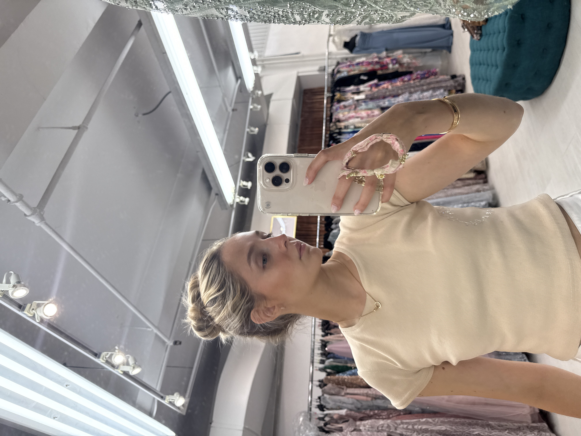 A blonde woman with her hair in a bun takes a mirror selfie in what appears to be a clothing boutique. She is wearing a cream-colored ribbed t-shirt and white pants. She is wearing a gold necklace and a gold bracelet. On her left wrist, she is wearing a bracelet with pink beads and charms. She is holding a white iPhone in her right hand. The background shows racks of dresses, and a teal ottoman. The lighting suggests an indoor setting, likely a retail space. The image is a close-up shot, focusing on the woman and her reflection. No specific New York City landmarks are visible.