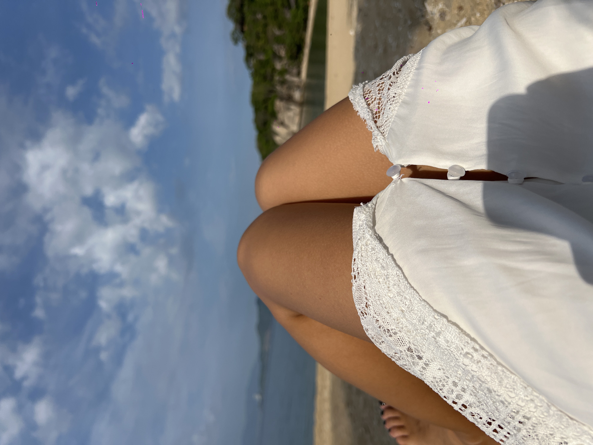 The image shows a first-person perspective of a person sitting on a sandy incline, looking out at a body of water with a hilly, tree-covered landscape in the background. The sky above is blue with scattered white clouds. The person is wearing a white top with lace trim and has tanned legs visible. The overall scene suggests a relaxing day at a beach or scenic overlook. The location is identified as Dong Ninh Hoa, Vietnam.