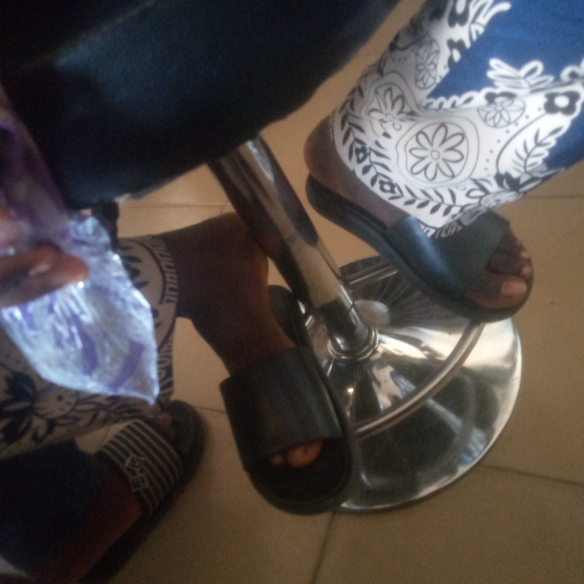 The image depicts two individuals seated indoors, viewed from a high angle focusing on their lower bodies. Both individuals have dark skin tones and are wearing open-toed sandals or slides. The person on the right is wearing blue pants or a skirt with a prominent white floral and leaf pattern and dark open-toed slides. Their feet are positioned on a shiny chrome footrest of a stool or chair. The person on the left is wearing dark pants and appears to have a striped pattern on their foot/sandal, consistent with slides. This individual is holding a translucent plastic bag containing purple material. The floor is light brown or beige, possibly tiled.