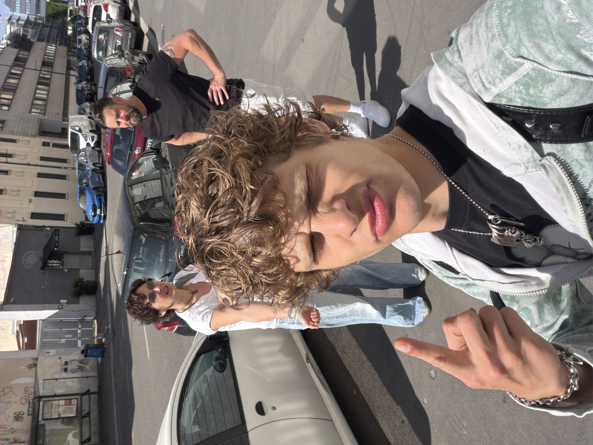 A photograph, taken in Milan, Italy, features a group of people on a city street lined with parked cars and buildings. The foreground subject is a young person with curly hair, wearing a black shirt, chain necklace, and a light-colored jacket. They are positioned at an angle, extending their hand towards the camera in a peace sign. Behind them and to the left, an older woman with short hair and sunglasses leans out of a white car, looking towards the foreground subject. Further back, another man with a beard and a tattoo on his arm stands with his hands on his hips, looking in the same direction. The background consists of parked cars along the street, with buildings on the left side, including a multi-story structure with numerous windows. Sunlight casts shadows on the ground.