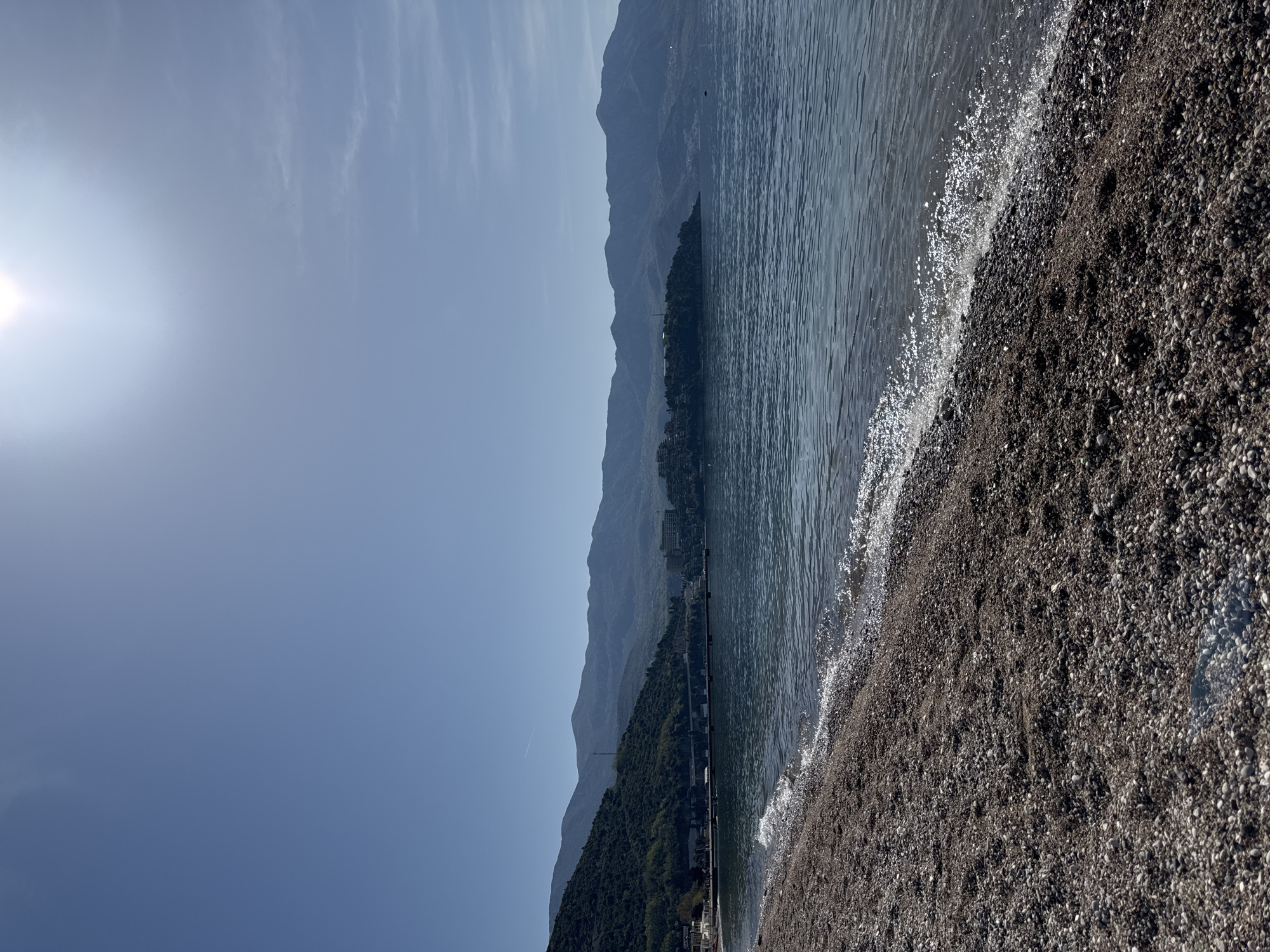 The image depicts a beach scene in Budva, Montenegro, characterized by a shingle beach meeting the Adriatic Sea. Mountains rise in the background under a clear sky with the sun visible. There are no people in the shot. The water has small waves and light reflections.