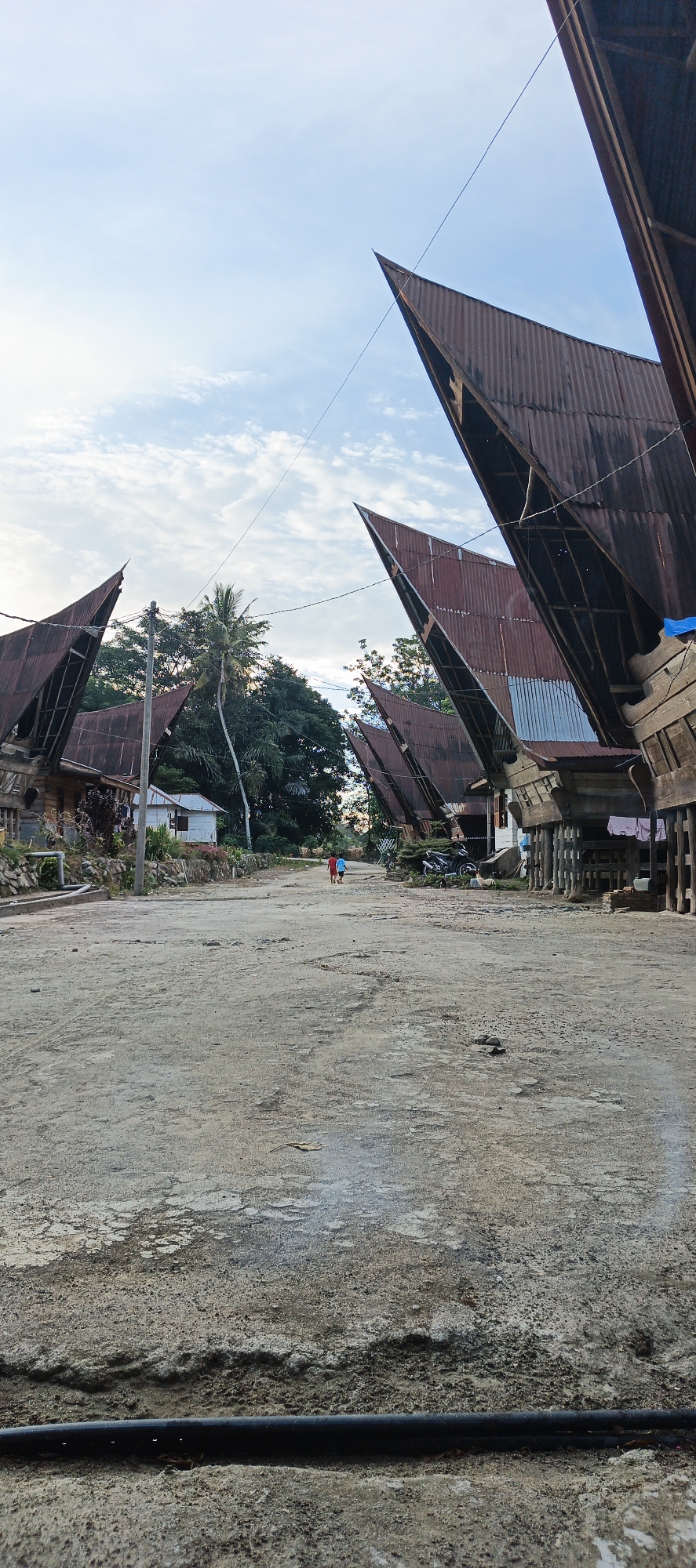 In Cinta Dame, Indonesia, two individuals, one wearing a red shirt and the other a blue shirt, walk away from the camera down a dirt road. They are positioned between traditional houses with distinct, upward-curving roofs constructed from corrugated metal. The houses are elevated on stilts. The environment is rural, with dense green trees visible in the background and a cloudy sky above. Laundry is hanging outside one of the houses. A motorcycle is parked near one of the houses. The foreground of the image shows a portion of the dirt road and a black pipe.