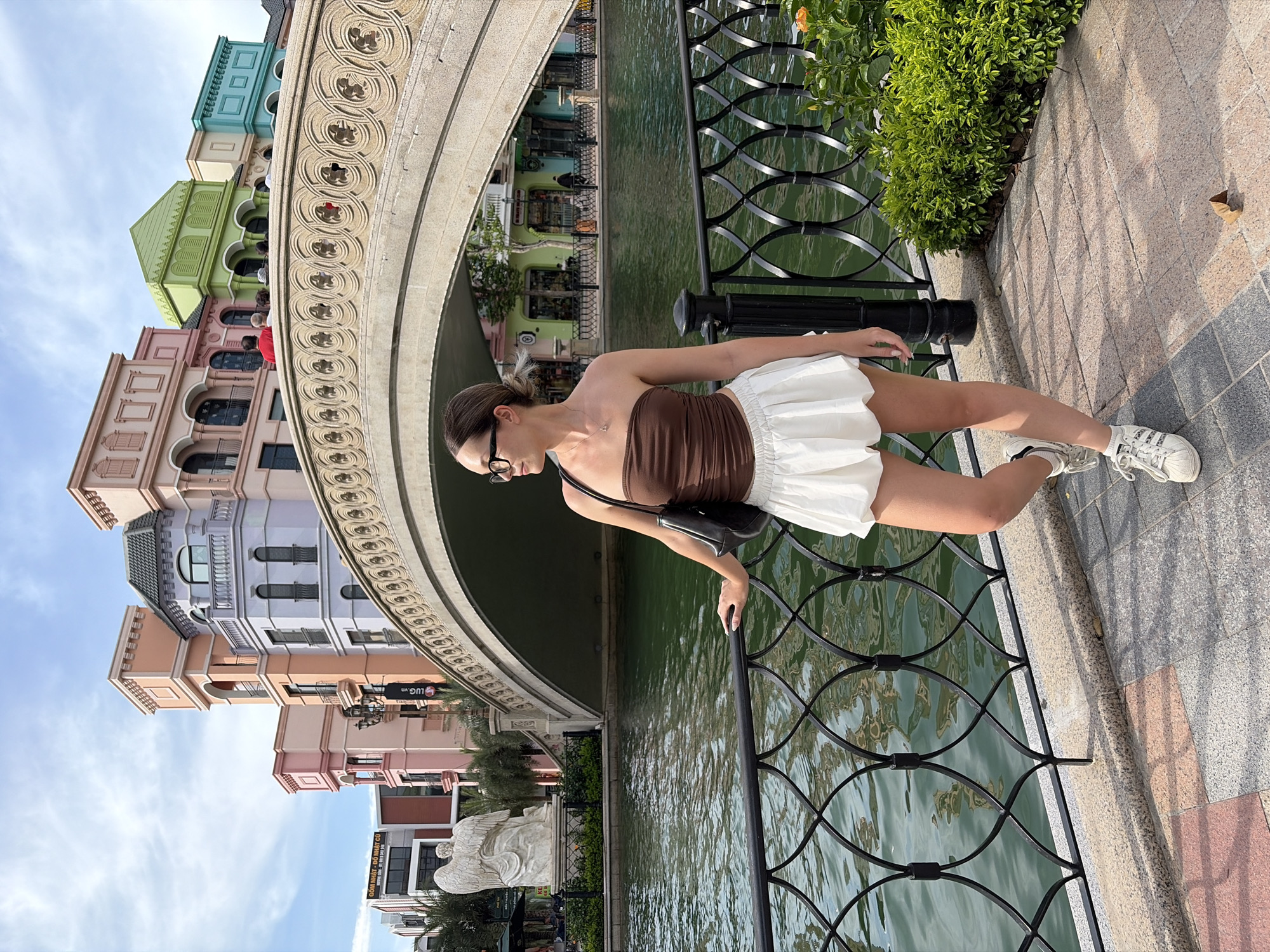 A young woman stands on a paved pathway next to a black metal railing, looking to her left. She has brown hair pulled back in a bun and wears glasses. Her outfit consists of a brown strapless top and a white ruffled skirt, with white sneakers. She is carrying a black purse over her right shoulder. Behind her, a body of water with green hues flows. A decorative stone bridge with intricate carvings spans across the water. On the far side of the water, a row of colorful buildings in pastel shades of pink, purple, orange, and green are visible. A large white statue of an angel stands near the water on the left. The scene is set outdoors under a partly cloudy sky. The location is identified as Nghĩa Trụ Commune, Vietnam.