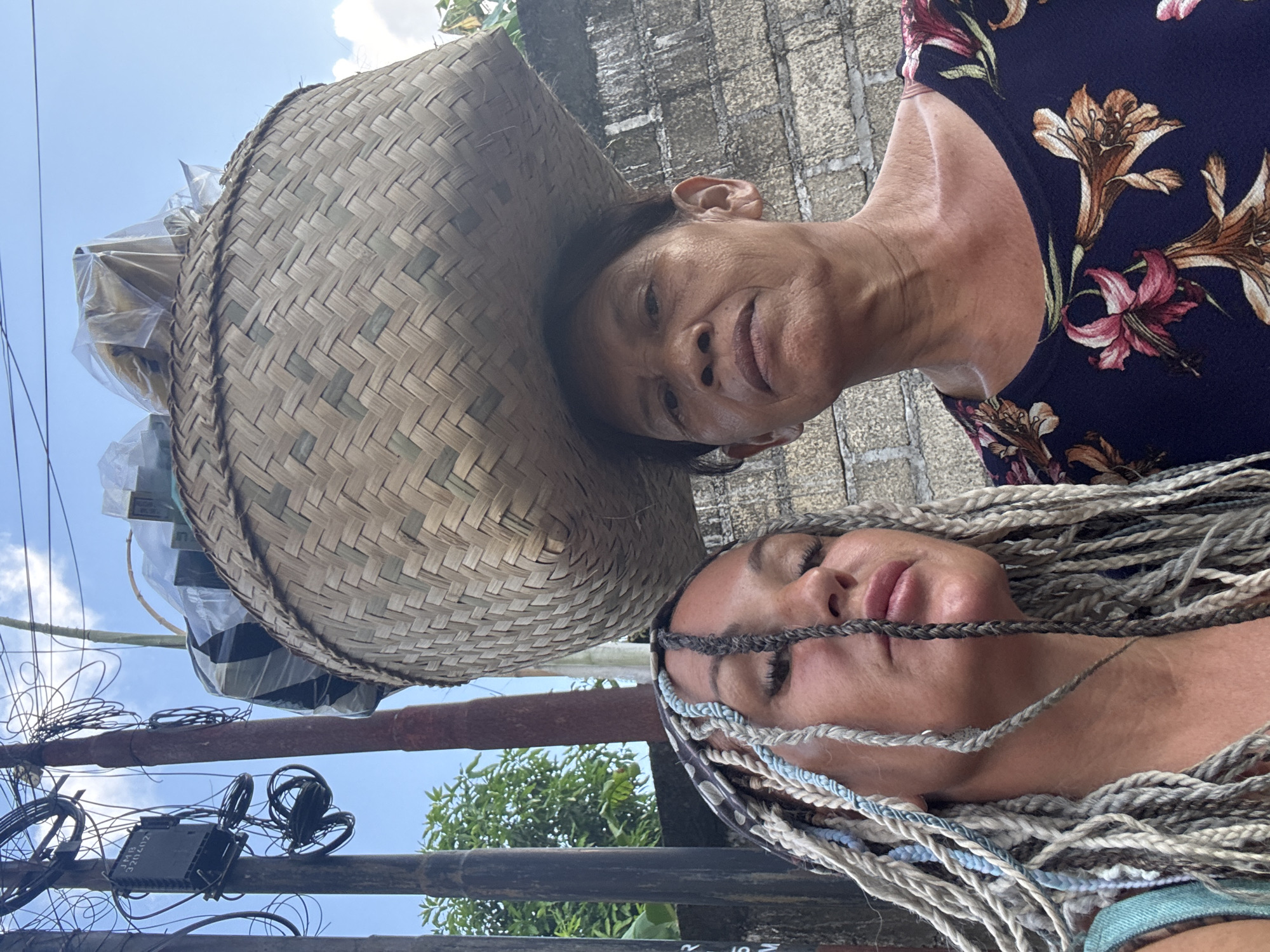 Two women stand close together. The first woman wears a floral top, a basket hat filled with goods and faces the camera. The second woman has braided hair and looks toward the camera. The picture was taken in the city of Kuta, Indonesia. The background includes a wall, trees, a utility pole with wires, and the sky.