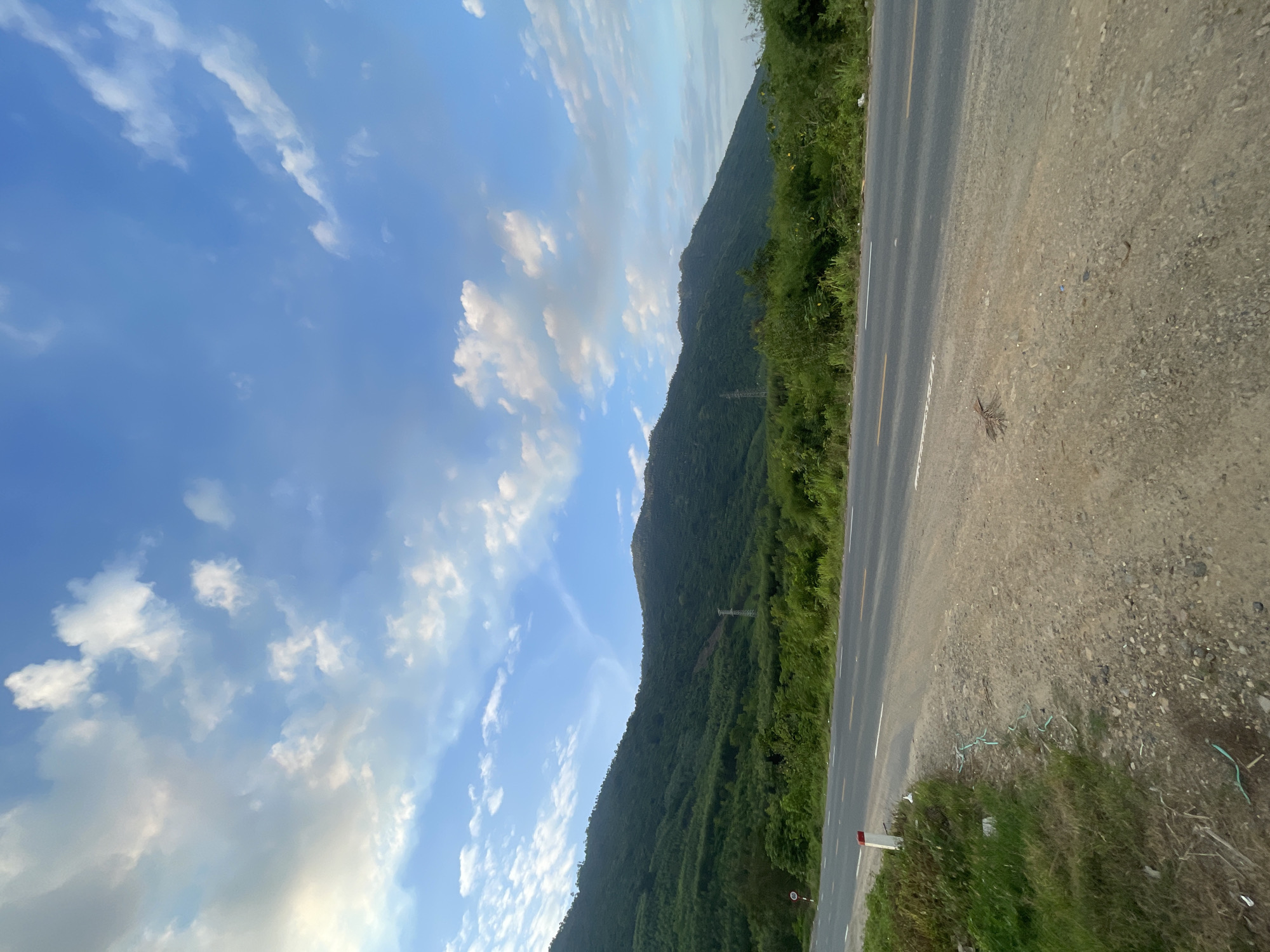 The image depicts a rural mountainous landscape in Tây Ninh Hòa, Vietnam, under a partly cloudy blue sky. In the foreground, a two-lane asphalt road with a double yellow center line and a solid white edge line runs horizontally across the frame. To the left of the road, a gravel and dirt shoulder transitions into a grassy verge containing scattered dry vegetation and a light green hose or pipe. A red and white reflective road marker post is visible on the left verge. The midground features dense, verdant mountains rising steeply, covered in lush green foliage. A prominent metal electricity transmission pylon is visible on a mountainside in the center-left, with a lighter, reddish-brown patch of earth on the slope nearby. The background consists of more distant mountains and a bright blue sky dotted with scattered white and light gray cumulus clouds. No people or vehicles are visible.