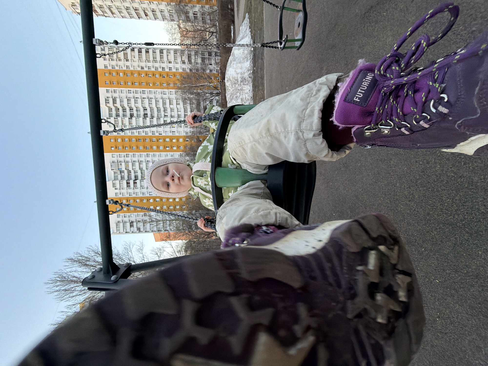A low-angle, close-up photograph depicts a young child seated in a green plastic swing on a playground. The child, estimated to be between 2 and 4 years old, wears a light pink knit hat, a green and white patterned jacket, and light beige pants, with a white lollipop or pacifier held in their mouth. Their hands grip the swing chains. In the immediate foreground, a blurred left foot and lower leg, belonging to the individual taking the photograph, are visible. This individual wears a dark, patterned pant leg and a purple hiking-style boot featuring the brand "FUTURINO" on its tongue, secured with purple and green speckled laces.
The playground surface is dark and appears to be rubberized. To the right of the child, an empty swing seat hangs from the same green metal frame. The background features several tall, multi-story residential buildings, some with prominent yellow-orange and white facades, indicative of urban residential architecture. Bare deciduous trees are visible between the playground and the buildings. A patch of melting snow lies on the ground behind the swings, suggesting late winter or early spring conditions. The sky is clear and pale blue, indicating daytime. This scene is located in a residential district of Moscow, Russia.