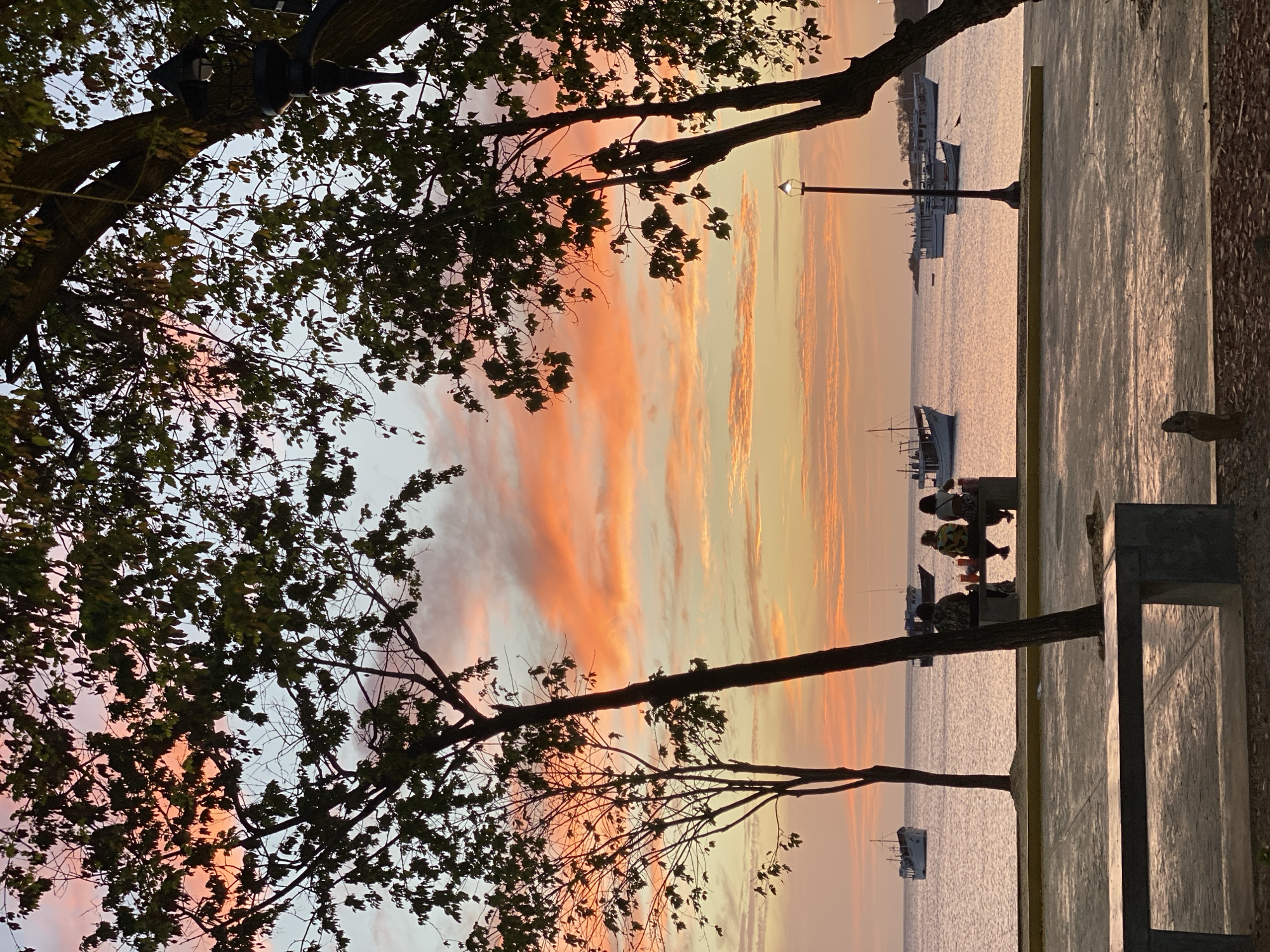 Captured from ground level, the image presents a tranquil sunset scene in Juan Griego, Venezuela. Silhouetted trees frame the upper left and right, providing a natural border to the vista. The sky is ablaze with a mix of orange and yellow hues as the sun sets. In the distance, boats dot the calm water. On the right, a concrete walkway extends into the scene, equipped with benches. People are seen sitting on these benches, observing the sunset, creating a peaceful tableau.