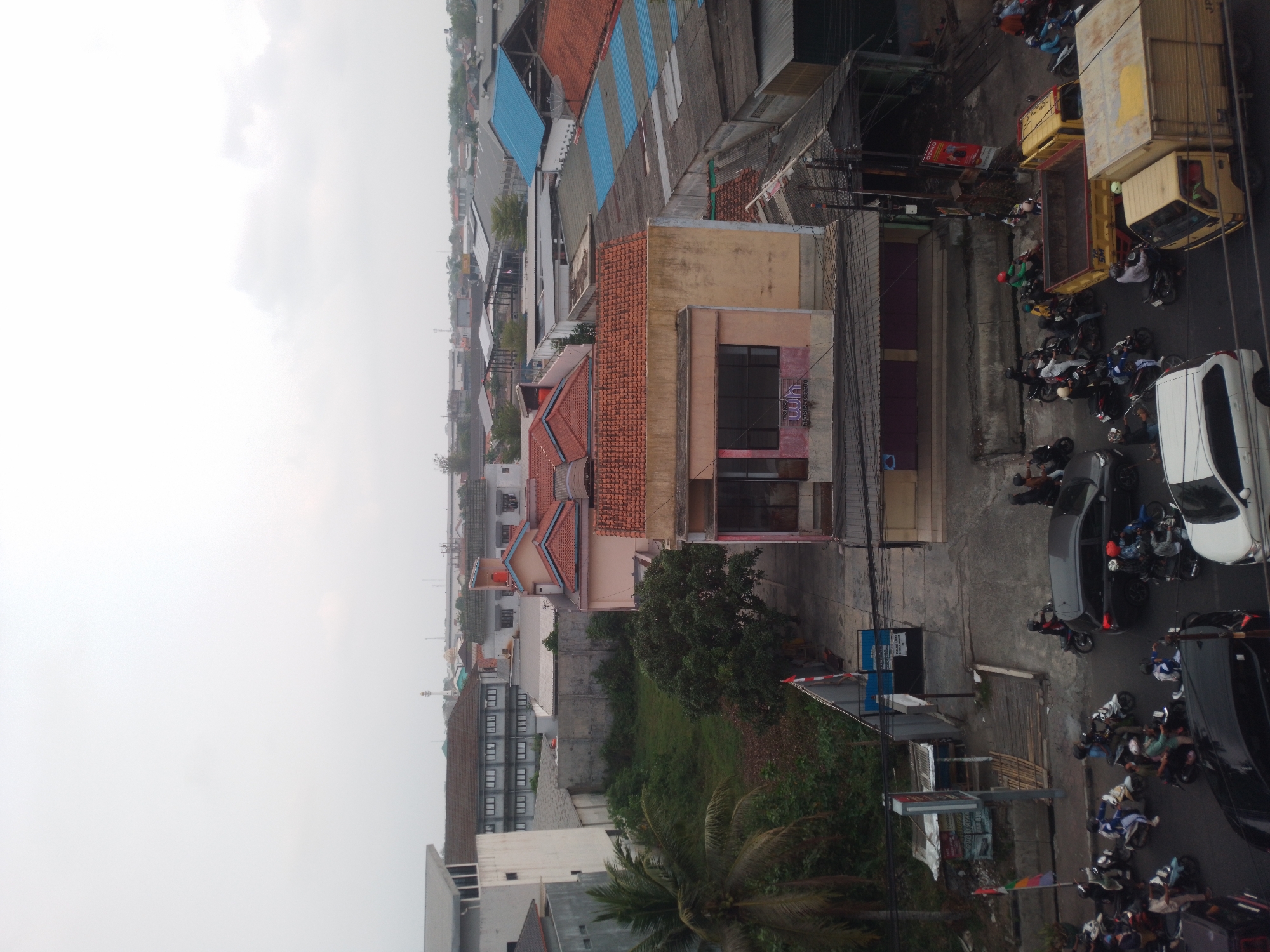 This is an overhead view of a street in Bumirejo, Indonesia, with buildings and traffic. The sky is overcast. In the foreground, several motorcycles and cars are visible, some in motion and some parked. A yellow truck is on the right side of the image. The buildings are a mix of residential and commercial structures, with tiled roofs and concrete walls. There is some greenery, including palm trees, in the middle ground. In the background, more buildings and industrial structures can be seen.