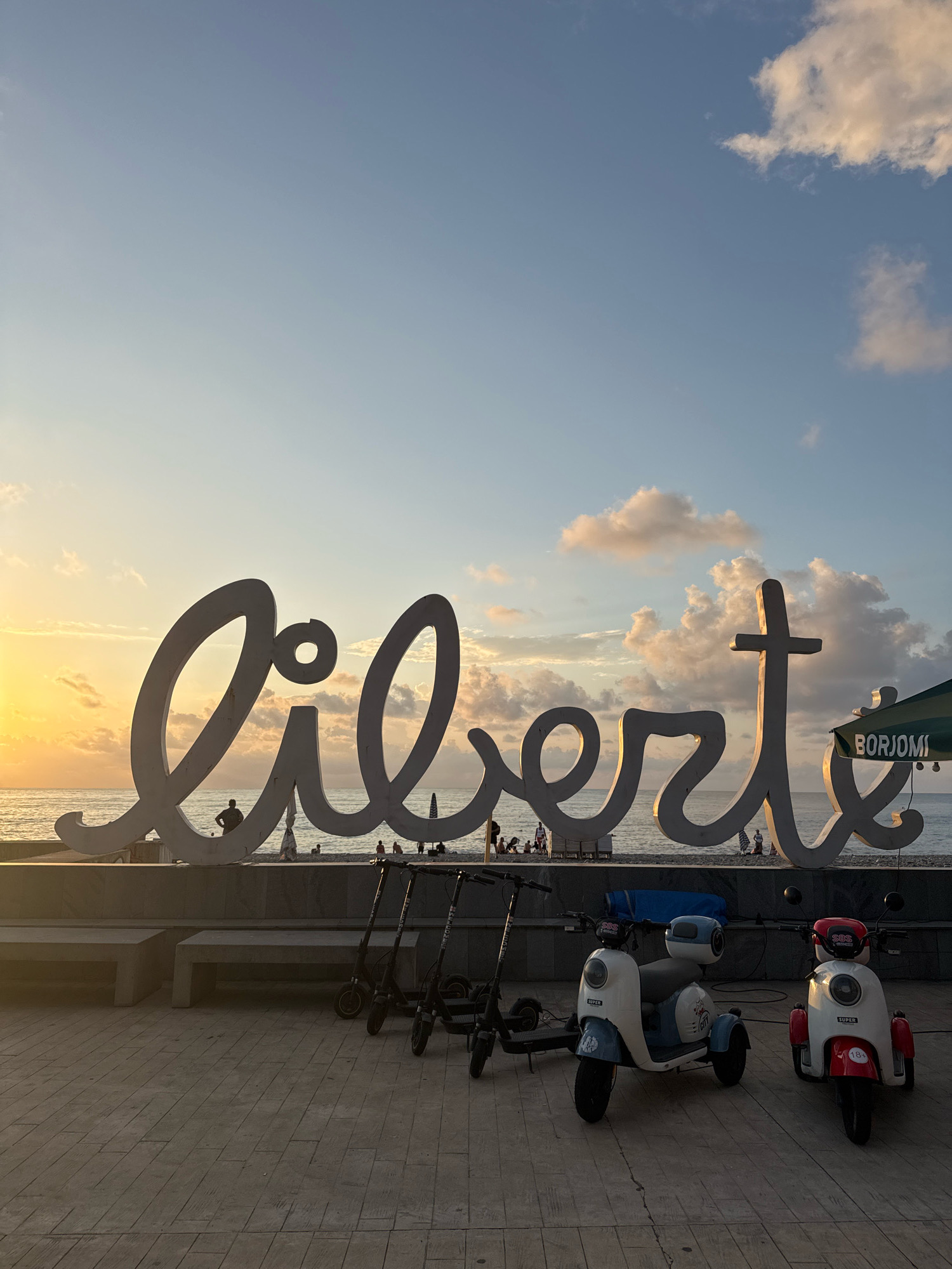 This is a vertical, medium shot photograph of the "Liberty" sculpture on Batumi Boulevard in Batumi, Georgia, during sunset. The large, white, cursive sculpture spelling "Liberty" dominates the center of the frame, with the Black Sea and a beach visible behind it. The sun is setting on the left, casting a warm glow on the sky and clouds. Several people are scattered on the beach and near the sculpture. In the foreground, three scooters are parked: two three-wheeled scooters, one white and blue and the other white and red, and one black electric scooter. The ground is paved with large, rectangular stones. A green umbrella with the "BORJOMI" logo is visible to the right of the sculpture. The sky is a soft blue with scattered, illuminated clouds.