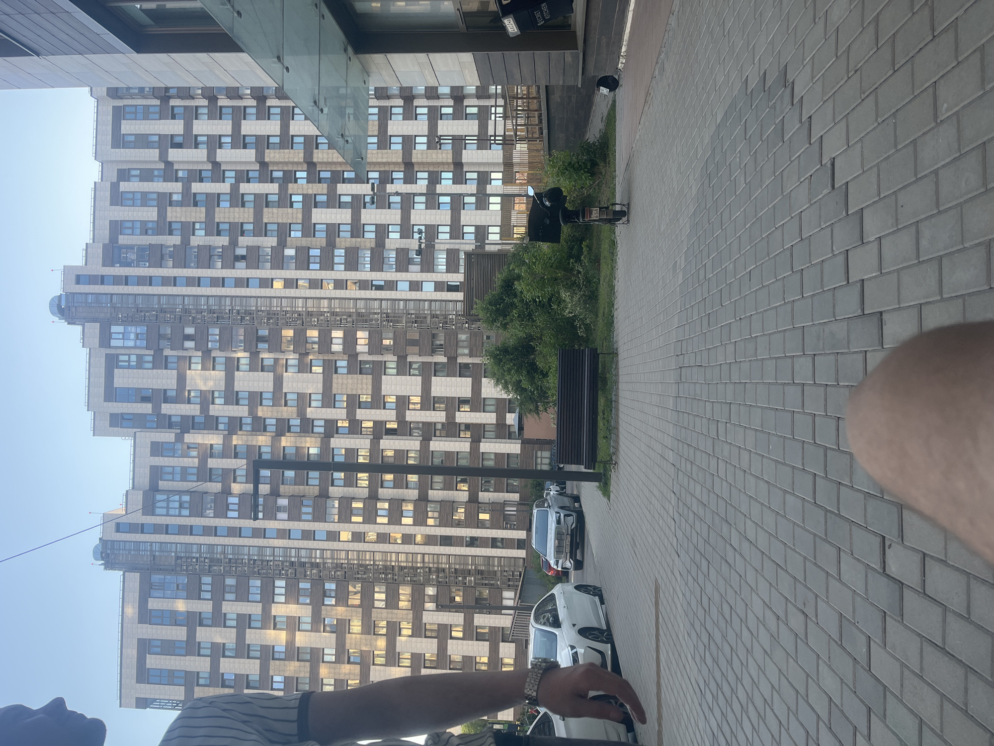 This is an outdoor, daytime photograph depicting a modern urban residential environment, identified as the city of Khimki, Russia. The perspective is low-angle, looking forward and slightly upwards from ground level.
In the immediate foreground, the left side of a person is visible, including the bicep, forearm, and hand of their left arm. They wear a light-colored, vertically striped short-sleeved shirt and a silver-toned wristwatch on their left wrist. Their hand is relaxed, fingers pointing downwards. In the bottom right corner, the lower portion of a person's leg or thigh is partially visible, indicating the camera operator's position. The ground is paved with rectangular grey interlocking pavers.
In the mid-ground, a paved street runs left to right, with parked vehicles. A white sedan is parked on the left, partially obscured by the foreground person. Further right, a white SUV with a visible Russian license plate "B 834 MM 799" (Moscow region) is parked. Behind it, a portion of a red vehicle is visible. Adjacent to the street, a strip of green landscaping features bushes and small trees. A black electric scooter with a large delivery box on its rear is parked on the paved area, facing away from the camera, displaying a partially visible license plate "65A". To its right, a dark slatted park bench is positioned. A dark, multi-bulb street lamp stands in the mid-ground, slightly right of center. Beyond the landscaping, a raised walkway or bridge structure with railings is discernible.
The background is dominated by a large, multi-story modern residential building with a distinctive facade composed of alternating light (beige/grey) and dark (brown/grey) panels in a rectangular pattern. Numerous windows reflect the daylight. A prominent external elevator shaft, enclosed in glass and metal, runs vertically up the center of the building. To the right, a darker building, likely commercial or part of the ground floor of the complex, features dark paneling and a large window display. A clear glass canopy extends over an entrance to this building. Two signs are affixed to the window of this building; one reads "SECRET KITCHEN", and another has the partial text "AX0346".
The scene is illuminated by natural daylight under a clear sky, with strong reflections off windows and vehicle surfaces. The architecture and vehicles are consistent with contemporary urban development in the Moscow region of Russia.