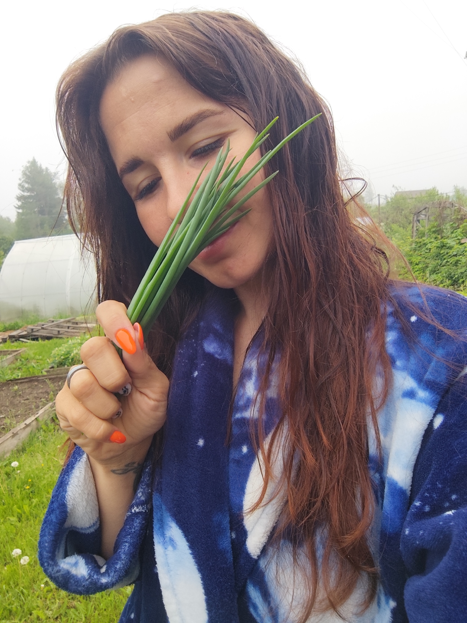 A woman with long, dark brown hair is captured in a close-up, outdoor shot, holding a bunch of green onions or chives to her face. Her eyes are mostly closed or looking downwards, conveying a sense of calm enjoyment or contemplation as she appears to smell the fresh greens. She is wearing a blue and white tie-dye or galaxy-print robe. Her left hand, adorned with bright orange nail polish and a ring, grasps the vegetables, with a faint tattoo visible on her forearm.
The setting is a garden or rural area in Фоки, Russia, characterized by visible garden beds and green grass in the mid-ground. A white, arched greenhouse or polytunnel is prominent in the background on the left, with distant trees and foliage further back. The sky is overcast and hazy, suggesting a foggy or damp day. The image captures a quiet, intimate moment, likely after harvesting fresh produce from the garden. No visible text is present.
