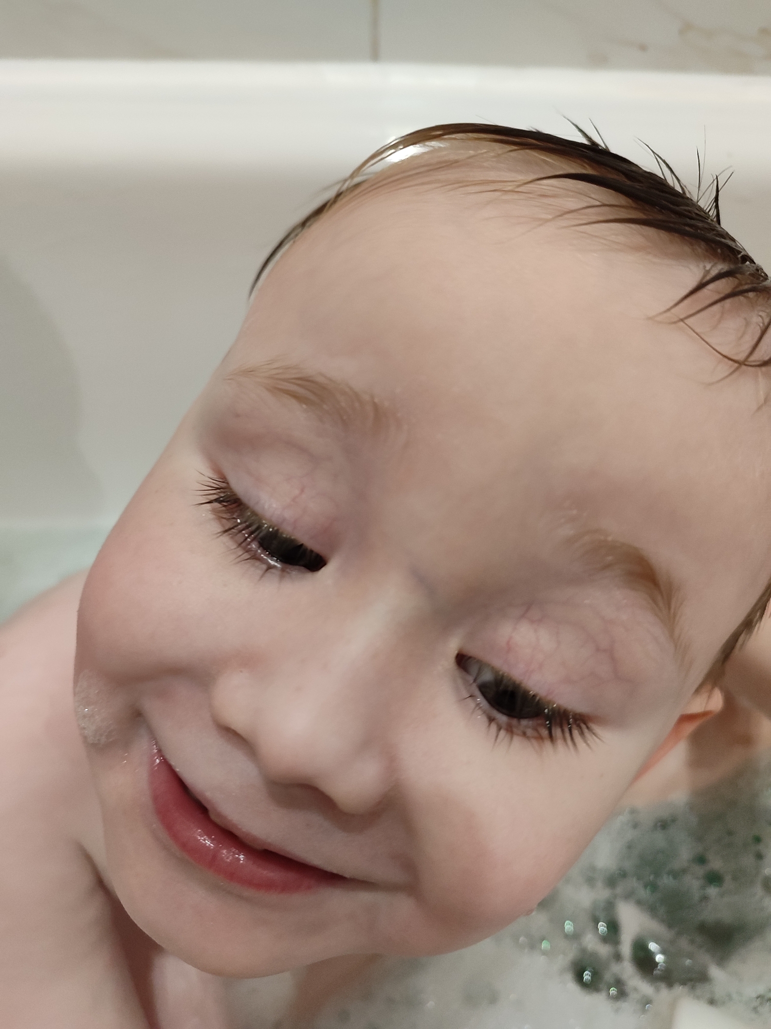 A close-up, high-angle photograph captures a young child, likely a baby or toddler, in a bathtub. The child's head and upper face dominate the frame, angled downwards and slightly to the left. The child has fair skin and wet, dark brown hair that is slightly spiky, indicating recent contact with water. A soft smile or grin is visible on the child's face, with rosy, glossy lips slightly parted. Bubbles from bath foam cling to the child's left cheek and around the mouth, and more foam is visible in the water in the bottom right of the image, appearing somewhat green due to reflections. The child's eyes are mostly closed or looking down, revealing prominent, long dark eyelashes. Delicate, fine veins are visible on the eyelids. The background shows the white interior of a bathtub and a white tiled wall above it, confirming an indoor bathroom setting. The overall emotion conveyed is one of joy and contentment during bath time. There is no visible text in the image. This scene takes place in Izhevsk, Russia.