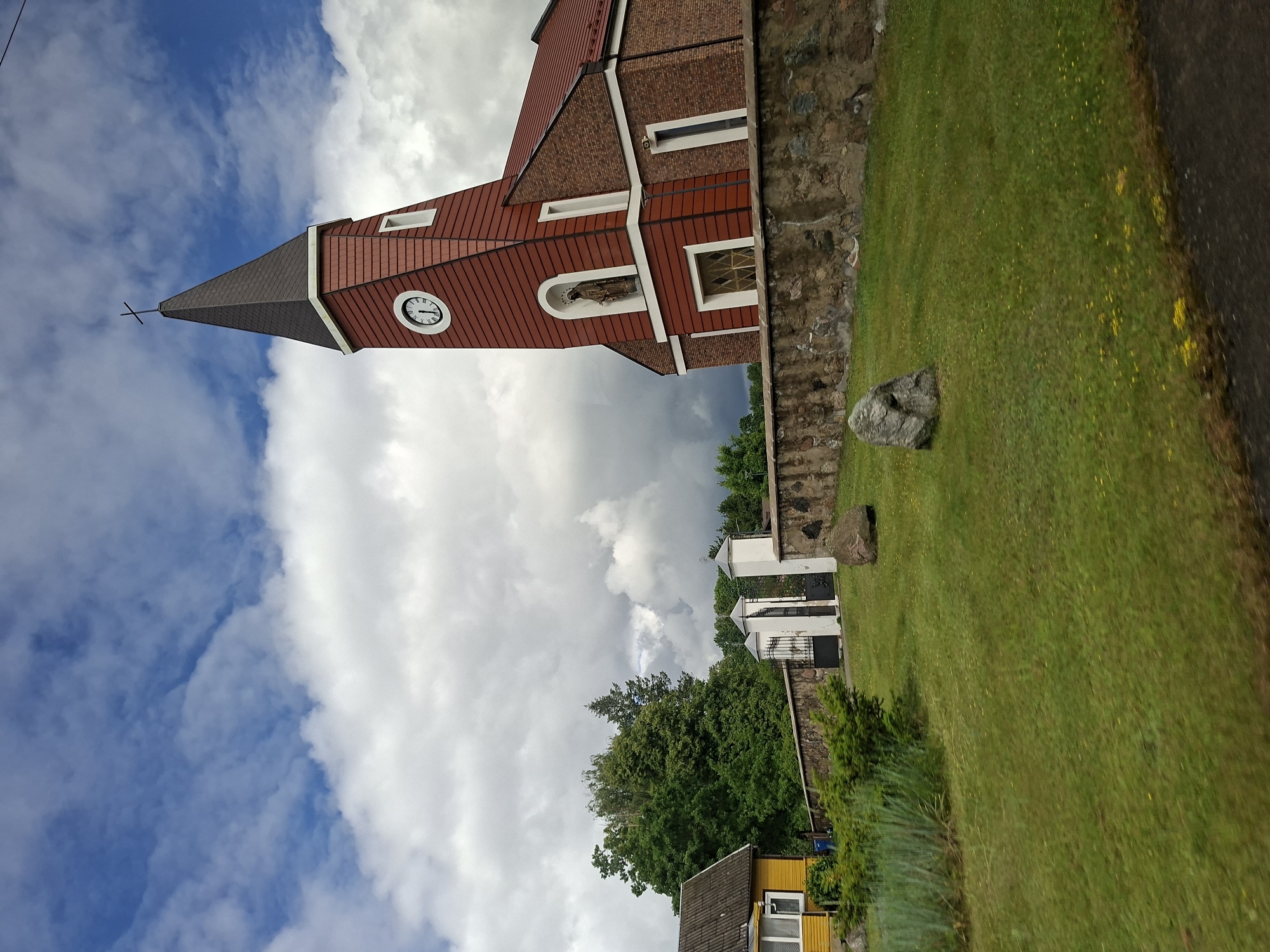 The image depicts the Church of St. Joseph in Balbieriškis, Lithuania. The church features a prominent red brick facade and a distinctive red tower with a dark roof, topped by a cross. A clock face is visible on the tower, along with a niche containing a statue. A stone wall with a gate separates the church grounds from the surrounding area, which includes a yellow house with a grey roof and green trees. The scene is set against a backdrop of a dynamic sky with both blue patches and large, white clouds, some of which appear dark and heavy, suggesting potential weather changes. The foreground is a lush green lawn with scattered yellow wildflowers and a few large rocks.