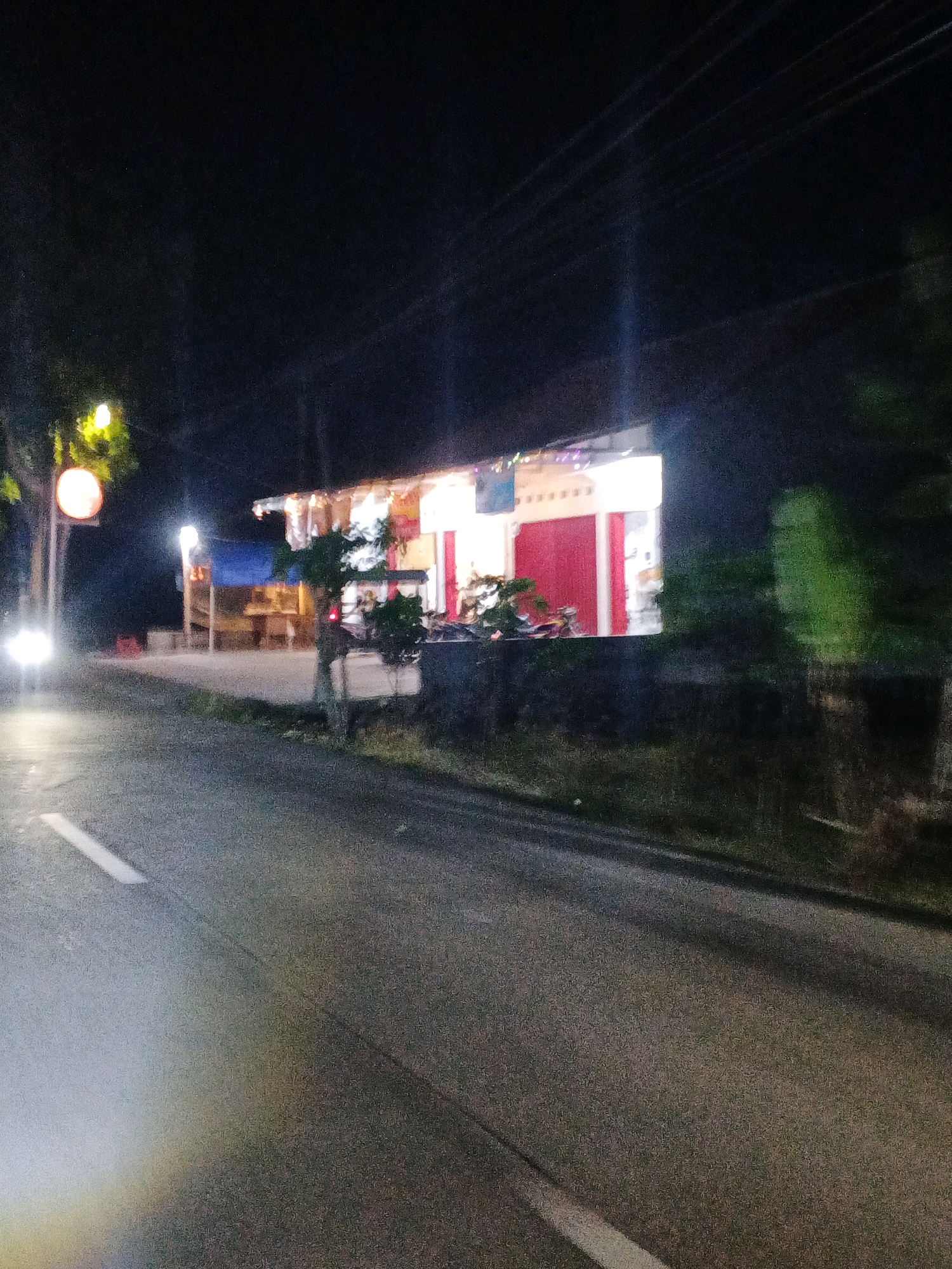 The image depicts a nighttime scene on a street in Planjan, Indonesia. A brightly lit building with red doors and white walls is visible on the right side of the road. The building appears to be a shop or restaurant, with decorative lights strung across its facade. A car with its headlights on is approaching from the left side of the image. Trees and foliage are present along the roadside, with some streetlights illuminating the area. The road surface is asphalt, with white markings indicating lanes. The overall atmosphere is one of a typical street scene in a semi-urban or rural Indonesian setting at night.
