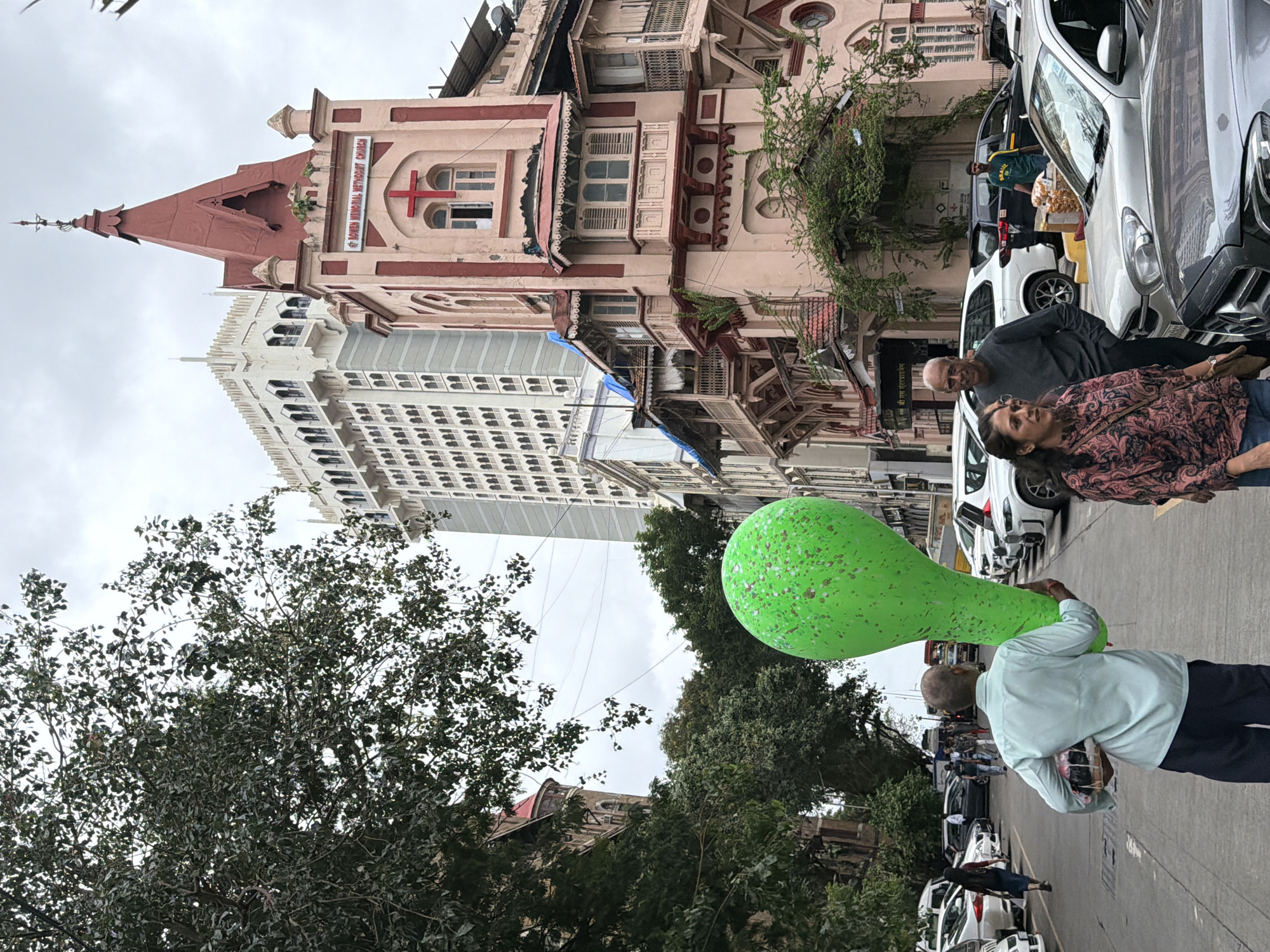 An overcast day in Mumbai, India, captured in a street-level photograph. A man walks away from the camera, carrying a large, green, speckled balloon and a basket of items in his left arm. He is dressed in a light blue collared shirt and dark trousers. To his right, a woman and a man are walking towards the camera, with the woman looking to her left and the man looking straight ahead. They are dressed in casual clothing. In the background, several cars are parked and driving on the street. A pink and red church building with a red cross on its facade is visible on the right, with "ST. BOWEN MEMORIAL METHODIST CHURCH" written above the cross. To the left of the church, a tall, ornate white building, which appears to be the Taj Mahal Palace Hotel, stands prominently. Trees with dark green foliage are scattered throughout the scene, with some branches extending into the frame from the left. The sky is cloudy.