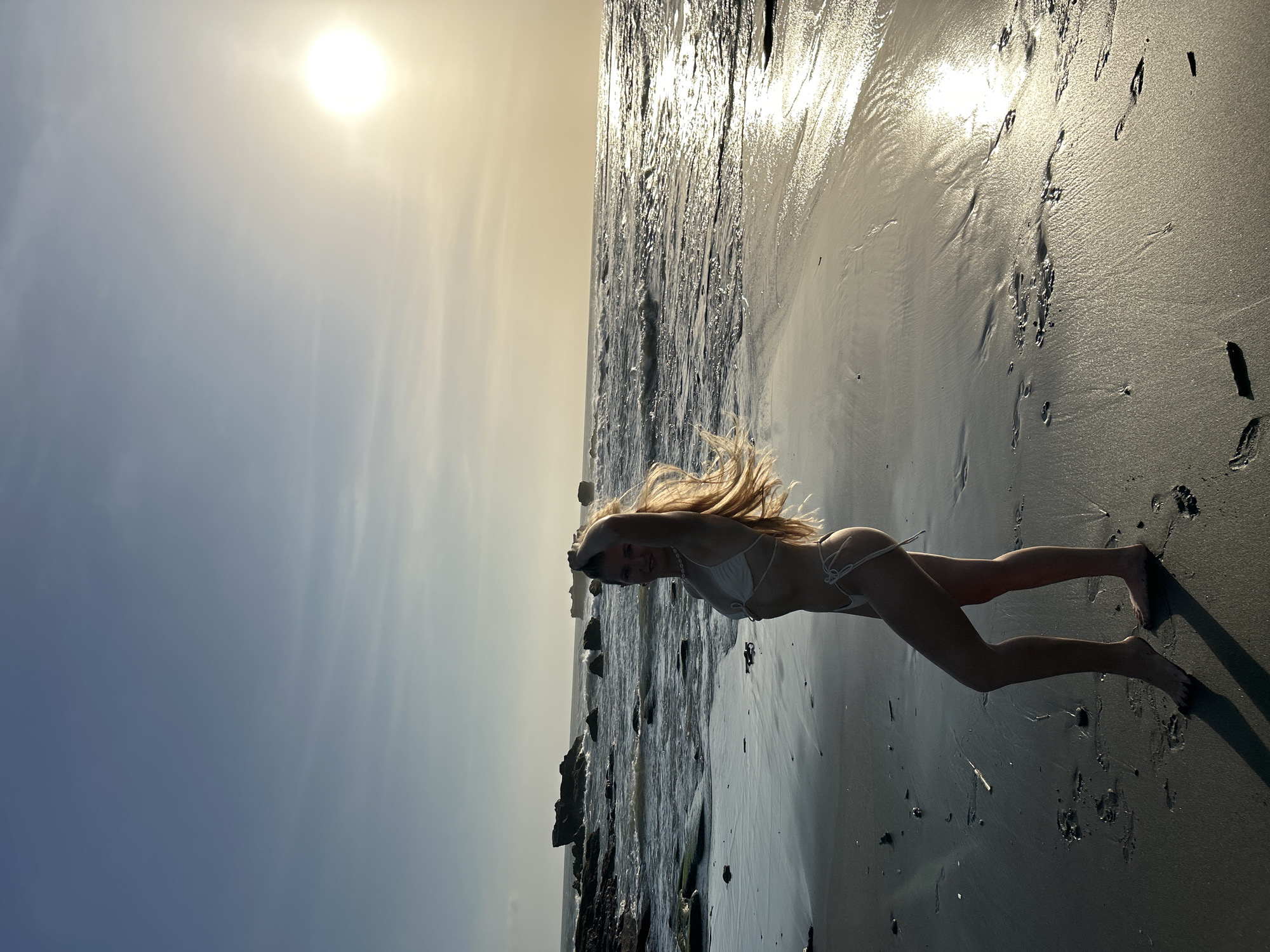 A woman in a white bikini lies on a beach. The background shows the ocean and the sun in the sky. There are rocks visible in the water. Footprints are in the sand. The location is likely on the beach somewhere in Mengwi, Indonesia.