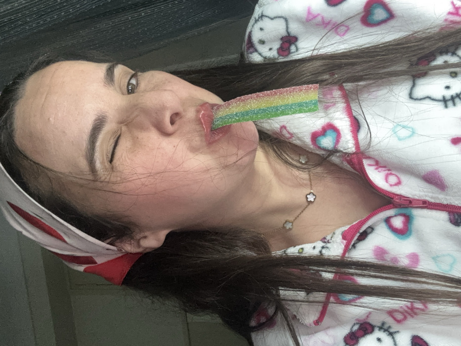 A woman with long brown hair and wearing a white headband and a Hello Kitty pajama top winks and holds a rainbow-colored candy strip in her mouth. She wears a delicate gold necklace with flower-shaped charms. Her skin is fair with rosy cheeks. The image is taken at an oblique angle.