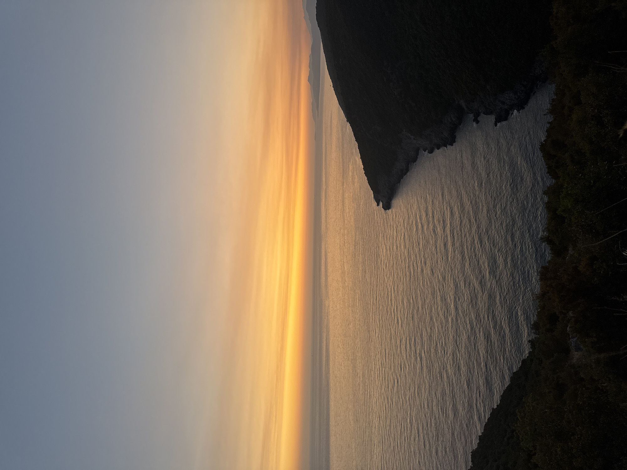 A vertical, high-angle shot captures a tranquil coastal sunset in Zagora, Montenegro. The sky transitions from a pale blue at the top to a vibrant, warm orange and yellow band near the horizon, with thin clouds streaking horizontally. Below the sky, the calm sea reflects the warm hues, its surface rippling gently. A dark, tree-covered hillside slopes down from the right foreground, meeting the sea at a rocky, indented coastline. The left foreground also shows a darker, vegetated hillside. In the distance, faint mountain silhouettes are visible on the horizon. The scene is devoid of human presence or any specific man-made objects, emphasizing the natural beauty of the landscape at dusk.