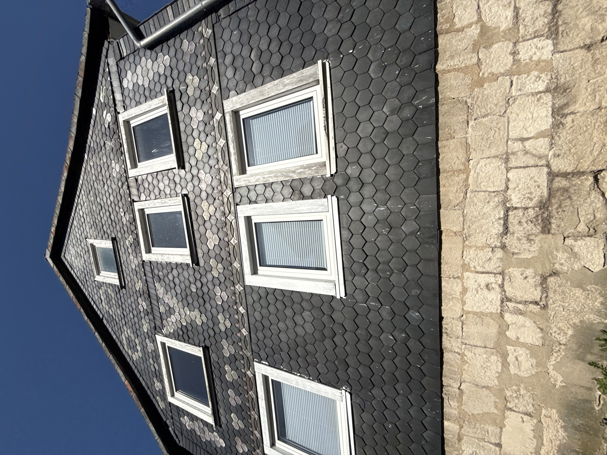 This is a low-angle, close-up shot of the upper portion of a building in Bad Sachsa, Germany, focusing on its roof and facade. The roof is covered in dark, hexagonal slate tiles. There are multiple windows visible on the sloped roof, appearing as dormers. The windows have white frames and are fitted with horizontal blinds. The facade of the building, below the slate tiles, is constructed of rough-hewn, light-colored stone blocks. The sky above is a clear, deep blue. There are no people or animals visible in the image. The scene depicts architectural details of a residential building.