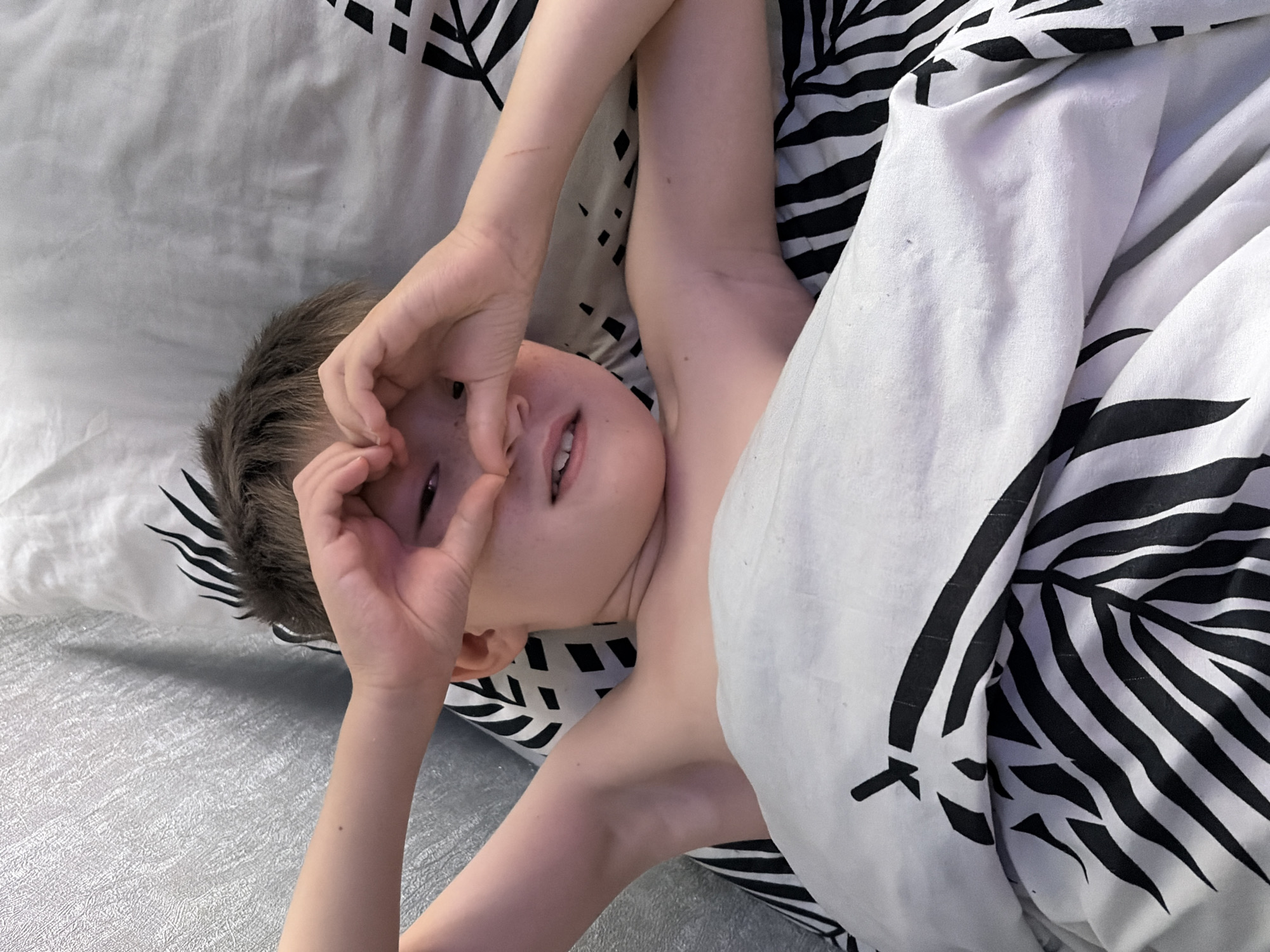 A young child lies in bed, partially covered by a duvet with a black and white palm leaf pattern. Their hands are forming a heart shape over their eyes. The child appears to be looking at the camera with a slight smile. The scene is indoors, likely a bedroom, with natural light. The overall mood is playful and endearing. There is no visible text in the image.