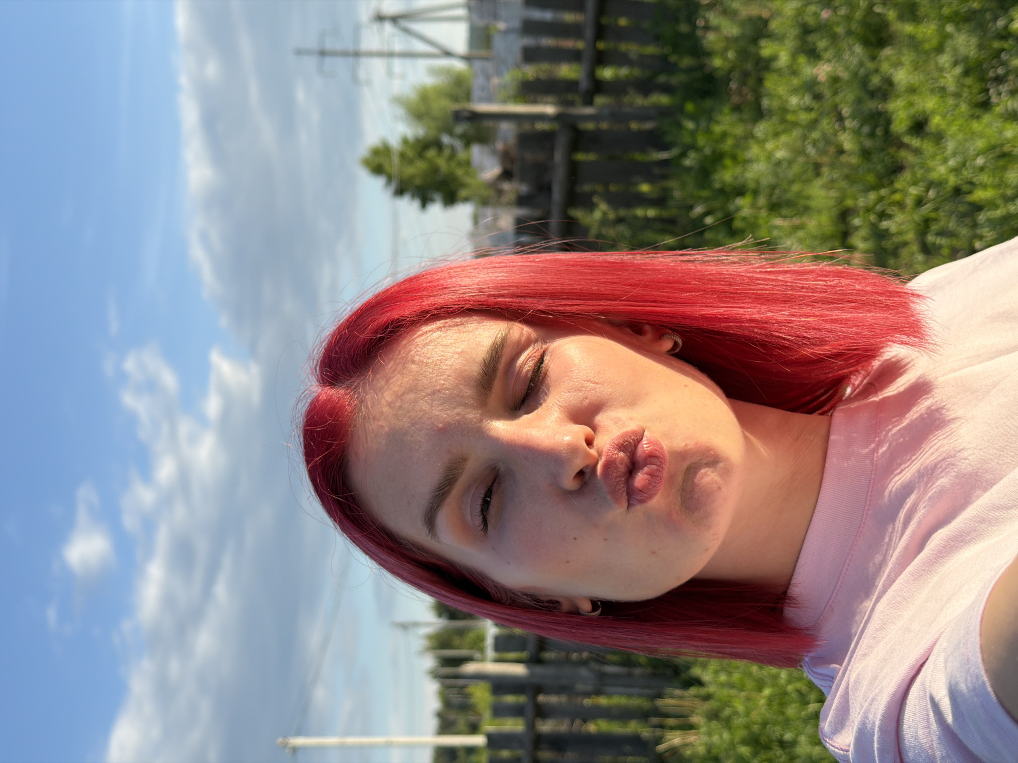 A female subject with bright red, shoulder-length hair is captured in a close-up selfie outdoors in Chaikovsky, Russia. She is wearing a light pink crew-neck t-shirt and a small hoop earring in her visible left ear. Her lips are pursed in a kissing gesture, and her eyes are partially closed, with the right eye (viewer's left) squinted, indicating exposure to bright sunlight. Strong sunlight illuminates the right side of her face, particularly her forehead and cheek. The background features a clear blue sky with scattered white clouds, a rustic wooden fence stretching horizontally, and green grass and foliage in the foreground and midground. Two utility poles with power lines are visible, one partially on the left and another more fully on the right, with distant green trees beyond the fence.