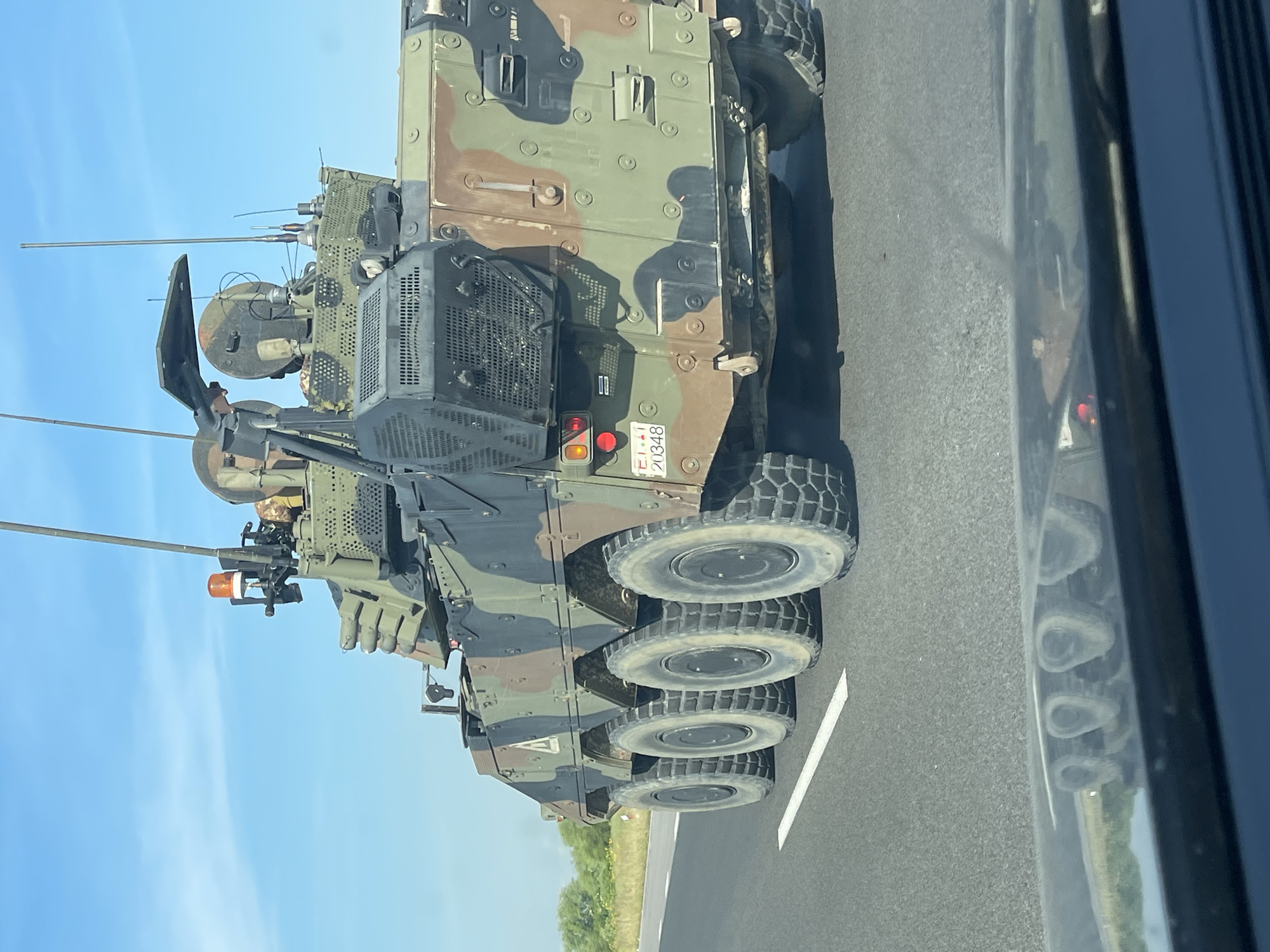 The media shows a military 8x8 wheeled armored vehicle, likely an Infantry Fighting Vehicle or Armored Personnel Carrier, traveling on a paved road. The vehicle is painted in a three-tone camouflage pattern (green, brown, and black). It is equipped with multiple antennas, suggesting advanced communication or electronic warfare capabilities. A remote weapon station or turret is mounted on top, featuring a visible gun barrel and an orange flashing light, possibly indicating its role in a convoy or warning. A soldier's head, wearing a helmet, is partially visible from a hatch near the weapon station.
The scene takes place during the daytime under a clear blue sky, suggesting good weather conditions. The road features a solid white line, indicating it could be a highway or main thoroughfare. Green foliage is visible in the background, to the left of the road. The photograph was taken from a civilian vehicle driving alongside, as indicated by the reflection in the right-side window.
A notable detail is the license plate located on the rear of the armored vehicle, which displays the text "E-141 20348". This format is consistent with Italian military vehicle registration. Given the provided location context of Загорци, Bulgaria, the presence of an Italian military vehicle suggests its involvement in a multinational exercise, deployment, or transport through the region.