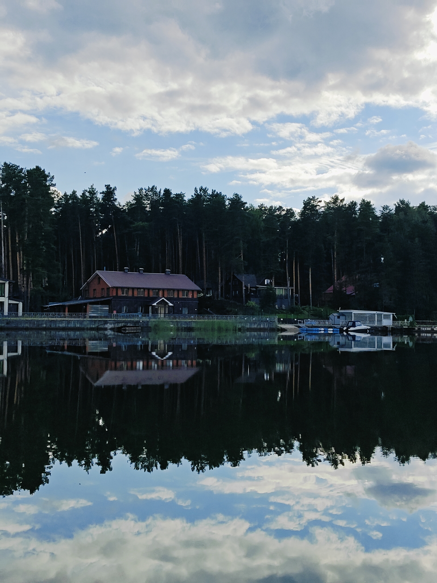 A vertical, medium shot captures a tranquil lake scene in Chaykovsky, Russia, on an overcast day. A dense, dark green forest lines the far bank, with tall, slender pine trees dominating the skyline. Nestled within the trees is a long, low-rise building with a reddish-brown brick facade and a darker tiled roof. A waterfront promenade with a metal railing extends along the edge of the property.
In the foreground, the calm surface of the lake acts as a mirror, reflecting the forest, the building, and the sky. The reflection of the building appears slightly distorted, with its windows and architectural details softened by the water. A few boats are visible docked near the shore on the right side of the frame. One is a sleek white and silver motorboat, and next to it is a blue inflatable boat.
The sky above is a mix of soft white and grey clouds against patches of pale blue, with the light diffused by the cloud cover. The overall atmosphere is serene and peaceful.