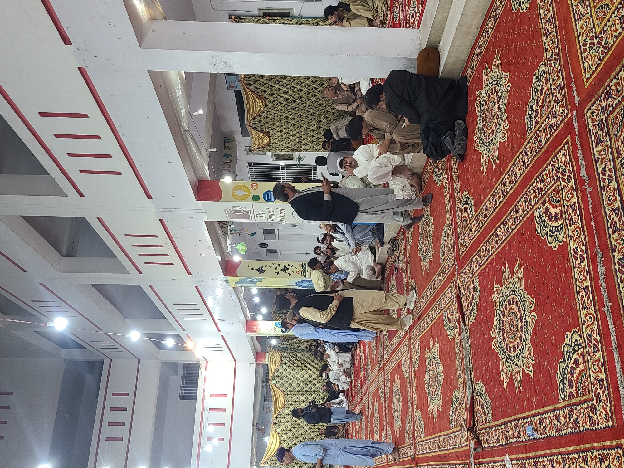 The image depicts an indoor gathering of approximately 30-40 adult males within a multi-story building in Quetta City Tehsil, Pakistan. The large space is illuminated by multiple exposed hanging light bulbs and features white walls with red vertical accent stripes and horizontal lines. The floor is covered with extensive red carpets adorned with intricate gold, black, and multi-colored patterns.
Numerous men, predominantly dressed in traditional shalwar kameez in varying shades of white, beige, light blue, and black, are present. The majority are seated on the carpeted floor or on low, raised platforms, conversing in small groups. Several individuals are observed looking at mobile phones. One man on the far left, wearing a light blue shalwar kameez and cap, stands facing right. In the middle-left, a man in a dark jacket and light blue shalwar kameez holds a video camera, seemingly recording the event. In the foreground-center, a man in a dark suit jacket and light gray pants stands holding a small object, possibly a phone. Another man near him, in a beige vest and shalwar kameez, holds a white bowl or tray. A large, patterned gold fabric drape covers a wall section in the background. A pillar on the right features a colorful mural with text partially visible as "The Smart School," indicating the venue is likely a school building. A barred window is visible on an upper level of the structure. The overall scene suggests a social or community gathering.