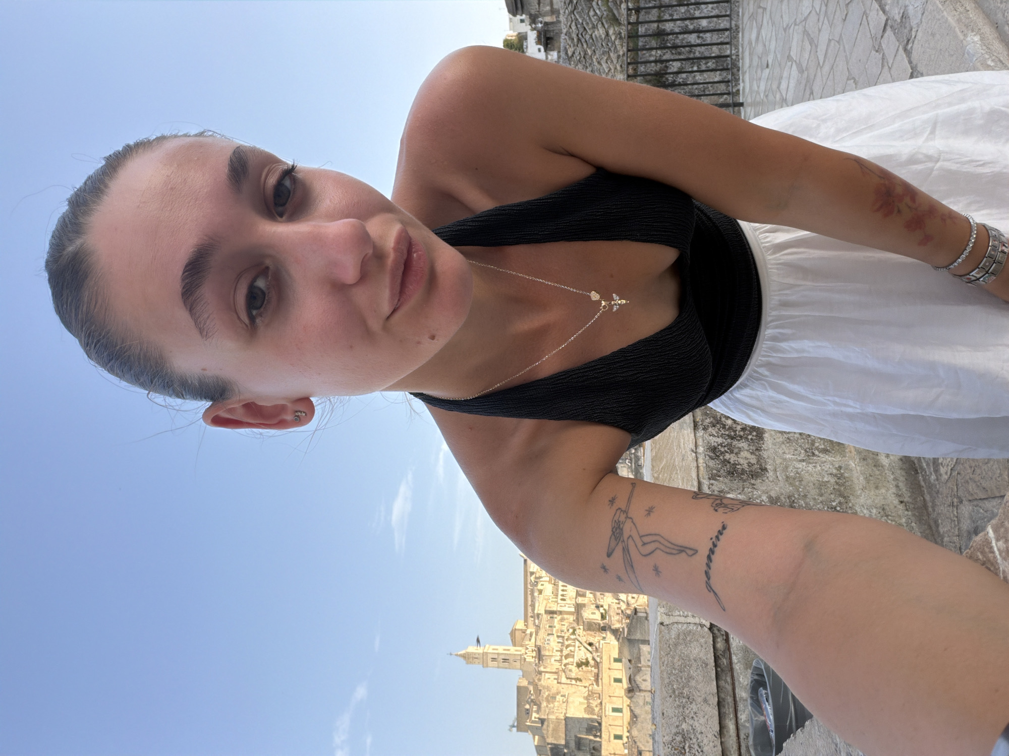 A young woman, with her hair pulled back into a ponytail, takes a selfie from a high vantage point. She is looking directly at the camera with a slight smile. She is wearing a black halter top and a white skirt. Around her neck, she has a silver necklace with a pendant of a bee and a small heart. On her left arm, she has two tattoos: one of a winged figure with stars and the word "gemini" written below it, and another on her right arm featuring red flowers.
In the background, the ancient city of Matera, Italy, is visible. The Sassi di Matera, a complex of cave dwellings carved into the rock, is a UNESCO World Heritage site. The city is characterized by its stone buildings, narrow streets, and steep staircases. A church tower rises above the cityscape. The sky is a clear, bright blue. The woman is standing on a stone walkway, with a metal railing to her right. A trash can is visible to her left, behind her.