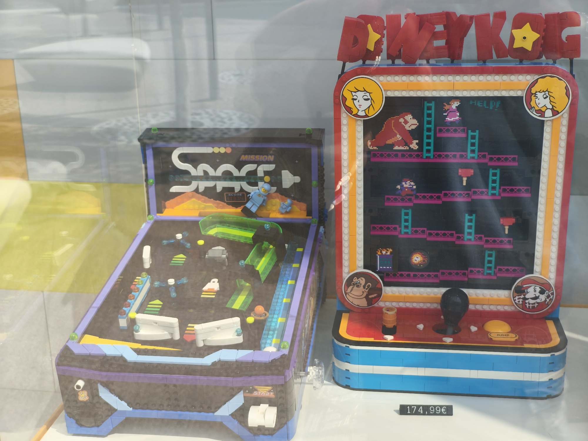A Lego set featuring a Donkey Kong arcade cabinet and a pinball machine is displayed behind a glass barrier in what appears to be a store. The Donkey Kong arcade cabinet is red with yellow accents and a pixelated representation of the game Donkey Kong on its screen. It has a joystick and buttons. The pinball machine is black and purple with a "SPACE MISSION" theme and a blue astronaut figure. A price tag of "174,99€" is visible in front of the display. The background is blurred and shows a tiled wall. The location is identified as Egaleo, Greece.