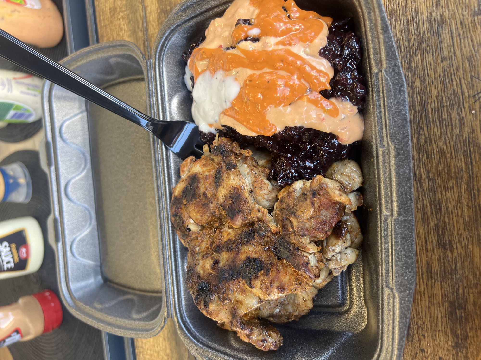 The image shows a takeout container of food, likely photographed on a wooden table, possibly in Burgas, Bulgaria (though this can't be definitively confirmed from the image alone). The container, made of black styrofoam, holds grilled chicken pieces with dark grill marks, dark rice, and a cream-colored and orange sauce drizzled on top of the rice. A black plastic fork rests on the edge of the container, partially inserted into the rice. A second, empty styrofoam container is next to it. A bottle of sauce is visible in the background.
