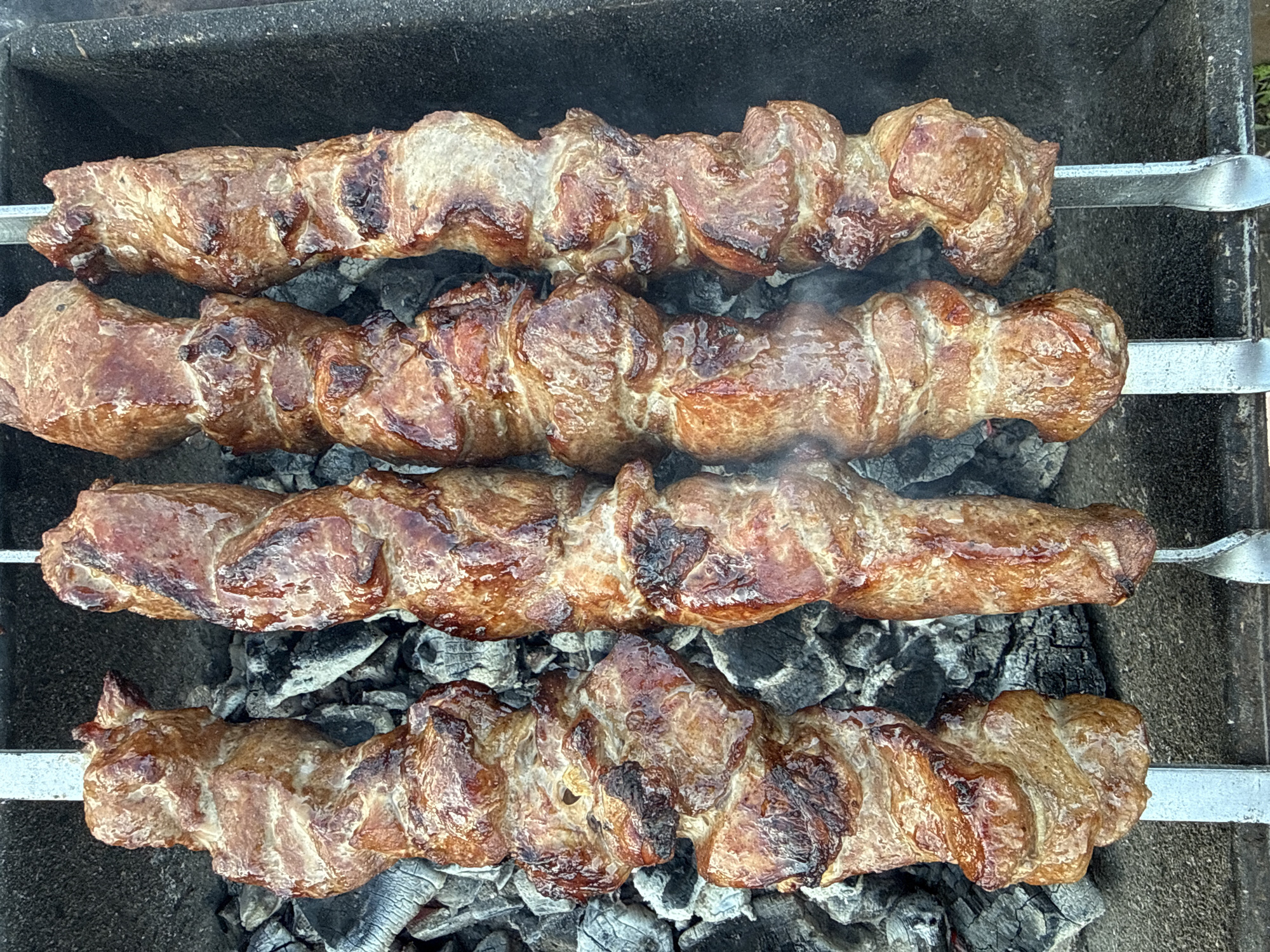 The image shows four metal skewers holding grilled meat, likely shish kebab, positioned over a bed of hot charcoal in a grill. The meat appears browned and cooked, with some areas darker than others. The grill's surface is visible around the charcoal. There is no indication of the location, Granovshchina, Russia, or the presence of any people.