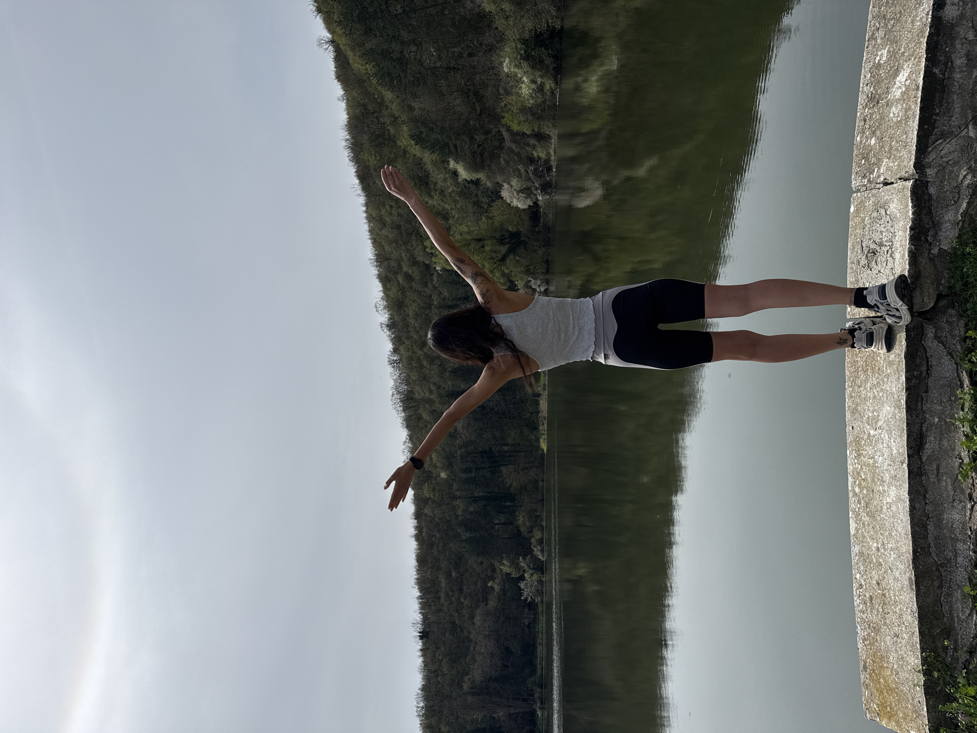 A young woman stands on a weathered concrete ledge. Her arms are outstretched, perpendicular to her body. She wears a light-colored tank top and dark athletic shorts and is facing away from the viewer. Her dark hair falls down her back. She also wears a wristwatch on her left wrist. A body of water spans the space directly behind her. This water reflects the image of a heavily forested hill. The sky is overcast. The location is near Pleven, Bulgaria, likely a dam or elevated walkway above the water.