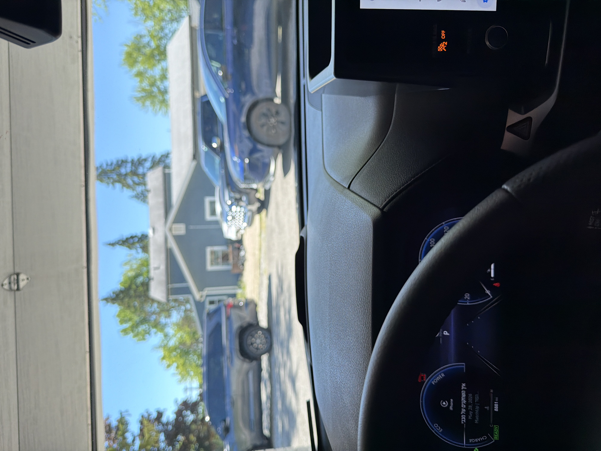 The image shows a view from inside a car, looking out through an open garage door onto a residential street. The car's dashboard is visible in the foreground, displaying a digital speedometer, battery charge indicators, and a date. Outside, a dark-colored SUV is parked in the driveway. Further down the street, two other vehicles are visible, a blue sedan and a light-colored pickup truck. A house with a blue facade and a gray roof is situated behind the vehicles. The sky is clear and blue, with green trees framing the scene. The overall impression is a sunny day in a suburban environment.