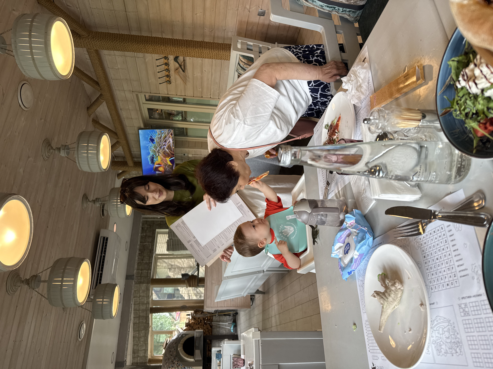 In a restaurant located in Sovkhoza imeni Lenina, Russia, a baby sits in a high chair, wearing a light blue bib and a red shirt. A woman in a green shirt is holding a menu and appears to be feeding the baby. To the right, an elderly woman in a white t-shirt and a dark patterned skirt leans in, also involved in the feeding interaction, holding a piece of food. The table in front of them is set with plates, cutlery, a bottle of water, and a packet of wet wipes. The restaurant has a rustic interior with wooden walls and ceiling, and decorative lighting fixtures. A television screen displaying fish is visible on the wall in the background.