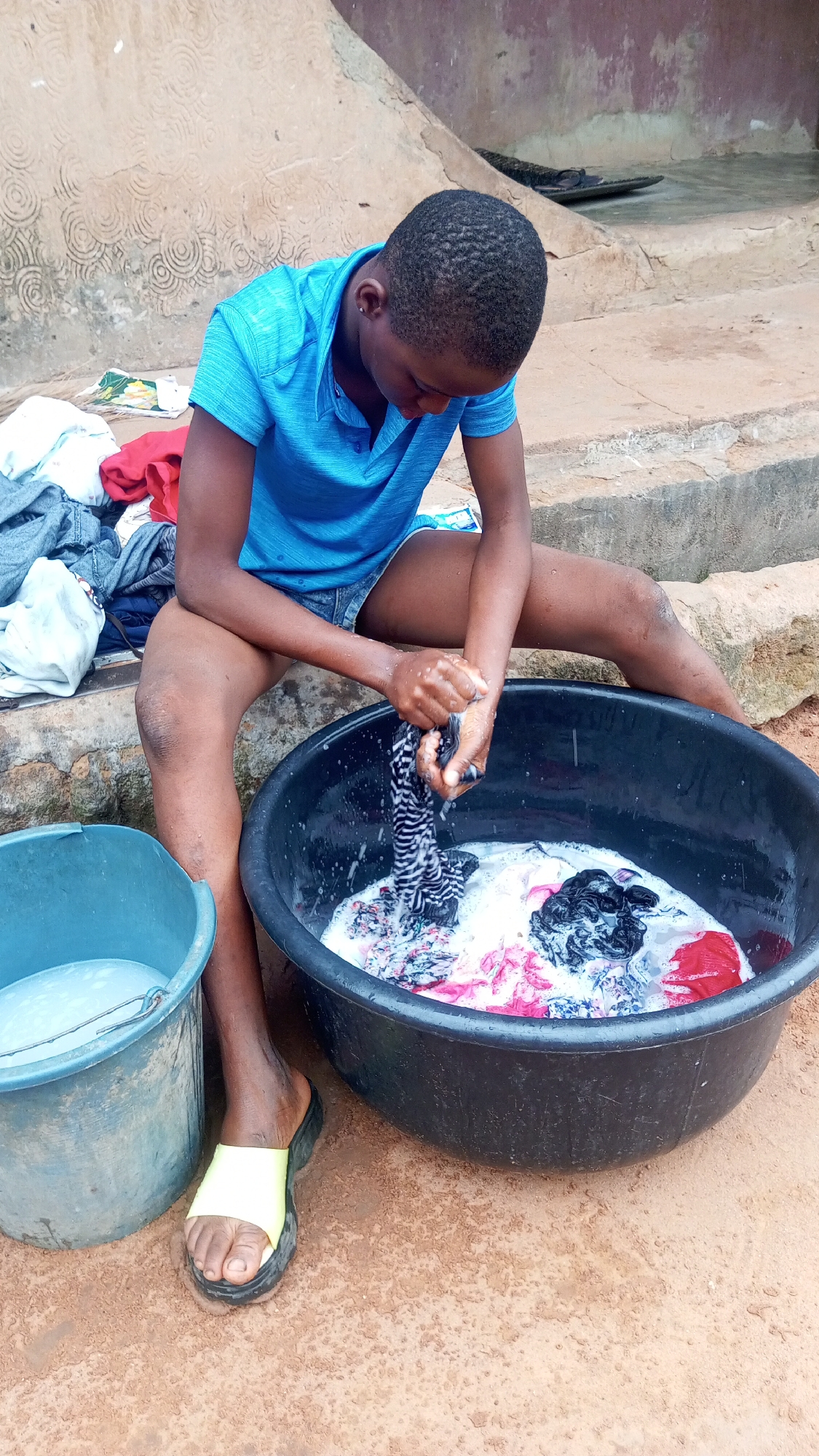 In Onitsha, Nigeria, a person wearing a blue shirt and denim shorts sits on a concrete ledge, washing clothes. They are perched with legs apart, a black basin filled with soapy water and colorful clothes between their thighs. The person's hands are submerged in the basin, wringing out a black and white striped garment. A blue bucket containing water is positioned to their left. They are wearing a yellow and black sandal on their left foot. Behind them is a wall with circular patterns etched into it. The ground appears to be dirt. Additional clothes are piled on the ledge near them.