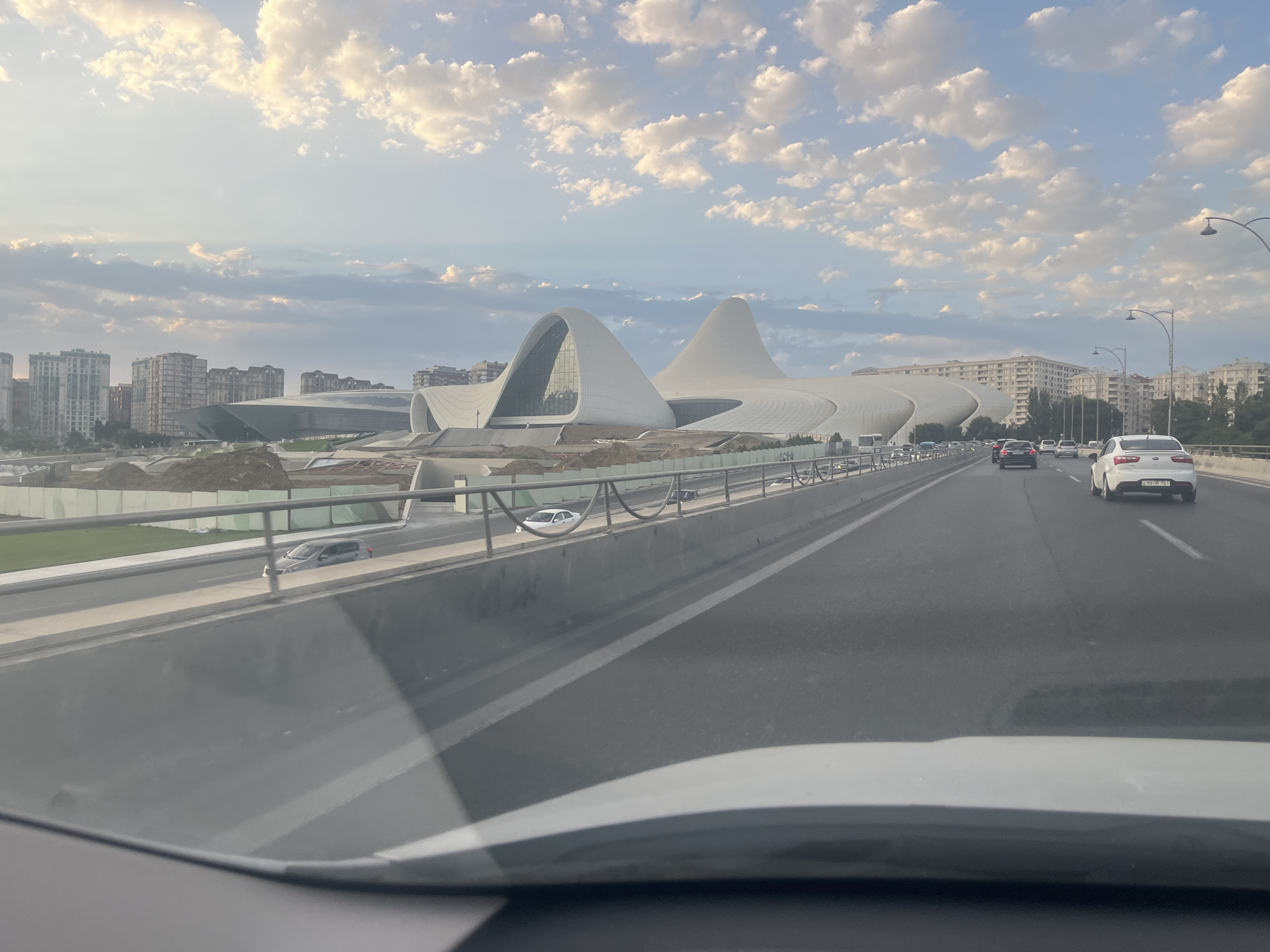 The image, captured from a moving vehicle, shows a view of the Heydar Aliyev Center in Baku, Azerbaijan. The iconic building, known for its flowing, wave-like architectural design, is prominently featured in the background against a sky filled with scattered clouds. In the foreground, a multi-lane highway is visible, with several cars driving on it. The highway is elevated, with a barrier separating it from a lower level where construction appears to be taking place, indicated by mounds of earth and temporary fencing. The overall scene depicts a blend of modern architecture, urban infrastructure, and ongoing development within the city.