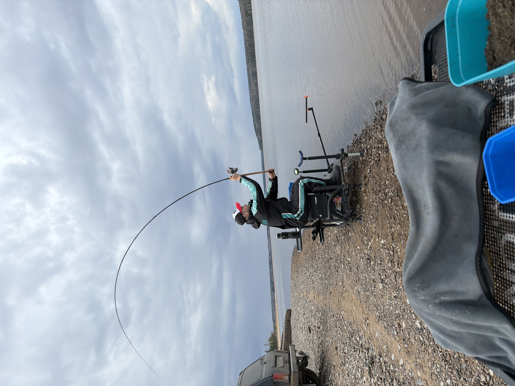 A man in a black and turquoise jacket and a red and white hat is fishing on the shoreline of a body of water under a cloudy sky. He is seated in a specialized fishing chair with attached accessories, including a rod rest and bait containers. His fishing rod is arched significantly, indicating he is actively reeling in a catch. The shoreline is gravelly and muddy, with a partial view of a vehicle in the lower left corner. The location is identified as the city of Chaikovsky, Russia.