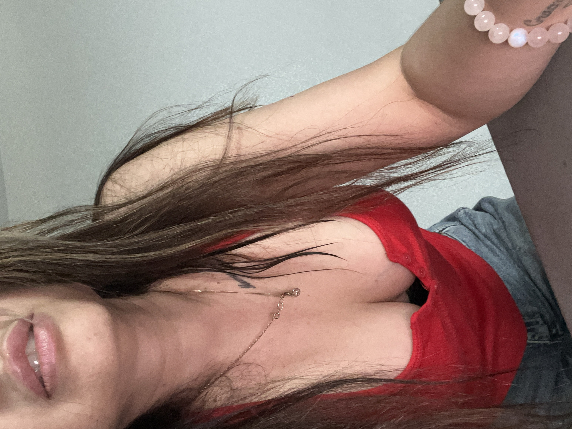 A close-up portrait shows a person, likely female, captured from an overhead, tilted angle. The person's long, dark brown hair cascades across their shoulders and chest. They are wearing a red top, possibly a tank top or bodysuit, and a glimpse of blue denim fabric, appearing to be shorts or jeans, is visible on the right side of the frame.
A delicate gold-colored necklace with a small pendant rests on their chest. On their right wrist, a bracelet made of light pink, spherical beads, some with an iridescent sheen, is visible. Part of the person's face, including their chin and slightly parted lips, is seen on the left. A small, dark tattoo, possibly a symbol, is visible on their upper chest, and another faint, illegible tattoo is on their right forearm. The background is a plain, light gray wall, suggesting an indoor setting. The image appears to be a casual selfie or self-portrait.
