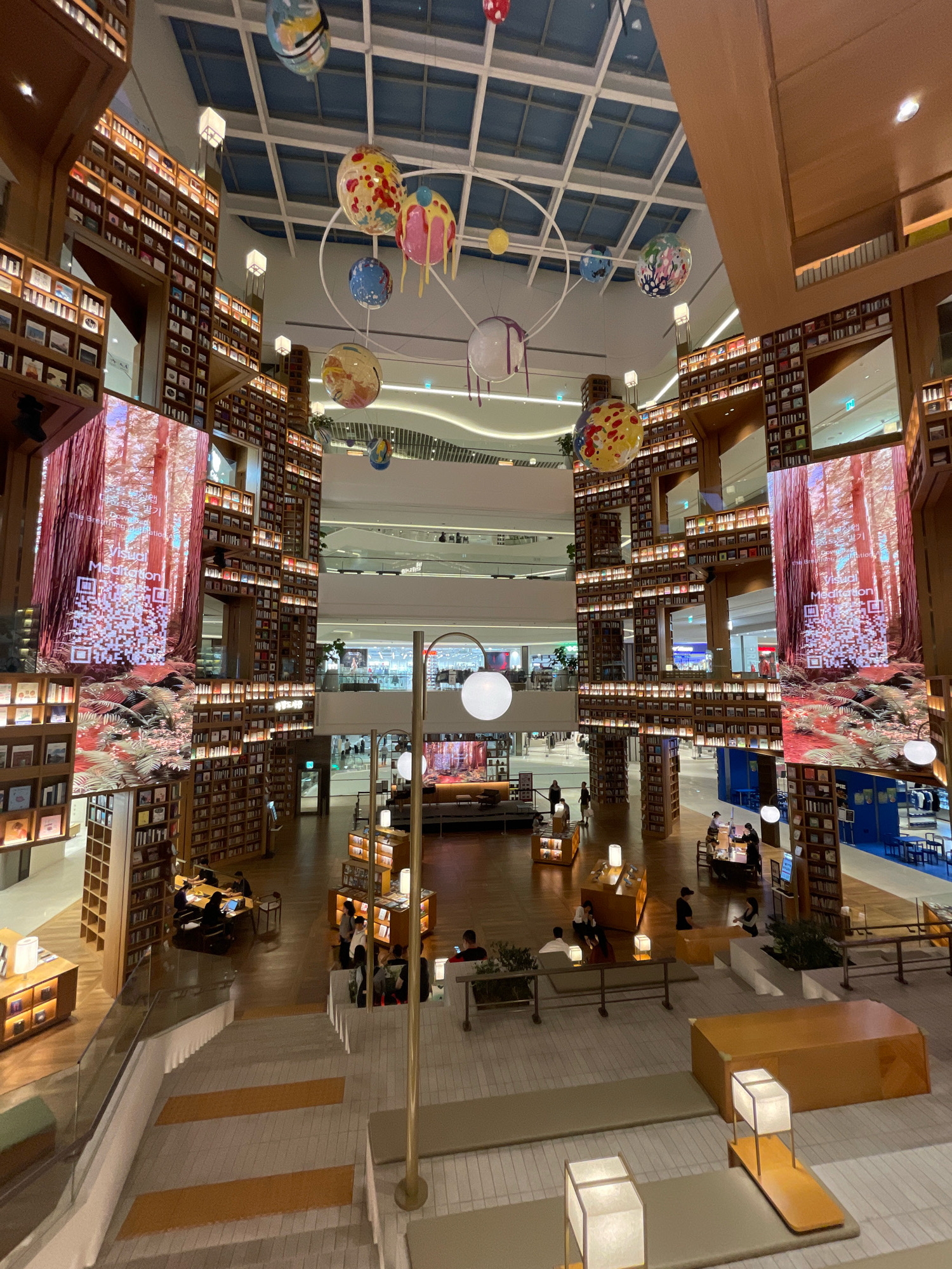 The image captures a multi-level interior space, likely a modern bookstore or library, with a high ceiling featuring a grid of skylights. The architecture is characterized by towering, floor-to-ceiling wooden bookshelves that wrap around open walkways and balconies on multiple levels. Suspended from the ceiling are several large, colorful spherical decorations that resemble planets or globes, some with dripping paint-like elements.
Large digital screens are strategically placed on the bookshelves, displaying immersive nature scenes, such as a forest with tall trees. One screen prominently features the text "Visual Meditation" in English, along with Korean script and a QR code.
The ground floor and intermediate levels are populated with people engaged in various activities. Some individuals are seated at tables, appearing to work on laptops or read. Others are standing and conversing. The space is furnished with wooden tables, chairs, and lounge-style seating areas. Modern lighting fixtures, including contemporary lamps with square shades, are visible throughout the scene. The flooring on the lower levels appears to be wood, while the visible stairs have a tiled surface with orange accent stripes. The overall ambiance is one of contemporary design and a focus on reading and relaxation. Based on the available information, this location is likely Starfield Library within the Starfield Hanam shopping mall in Hanam-si, Gyeonggi-do, South Korea, which is often associated with Suwon-si due to proximity and similar types of large-scale commercial developments in the region.
