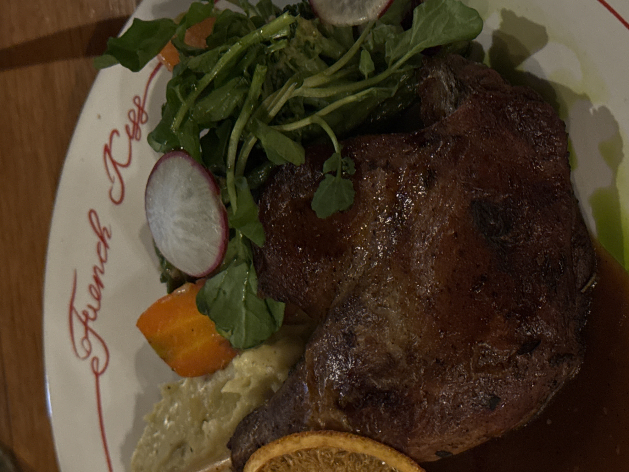 The image shows a close-up of a plate of food, likely at a restaurant. The plate has a red cursive inscription that reads "French Kiss". The food consists of a piece of roasted meat, possibly duck, served with mashed potatoes or a similar purée, a salad of leafy greens with radish slices and carrot. A slice of orange, likely candied or dried, is visible on the left side of the plate. The lighting suggests an indoor setting, possibly a dimly lit restaurant. There are no people visible in the image. The image's resolution is moderate.