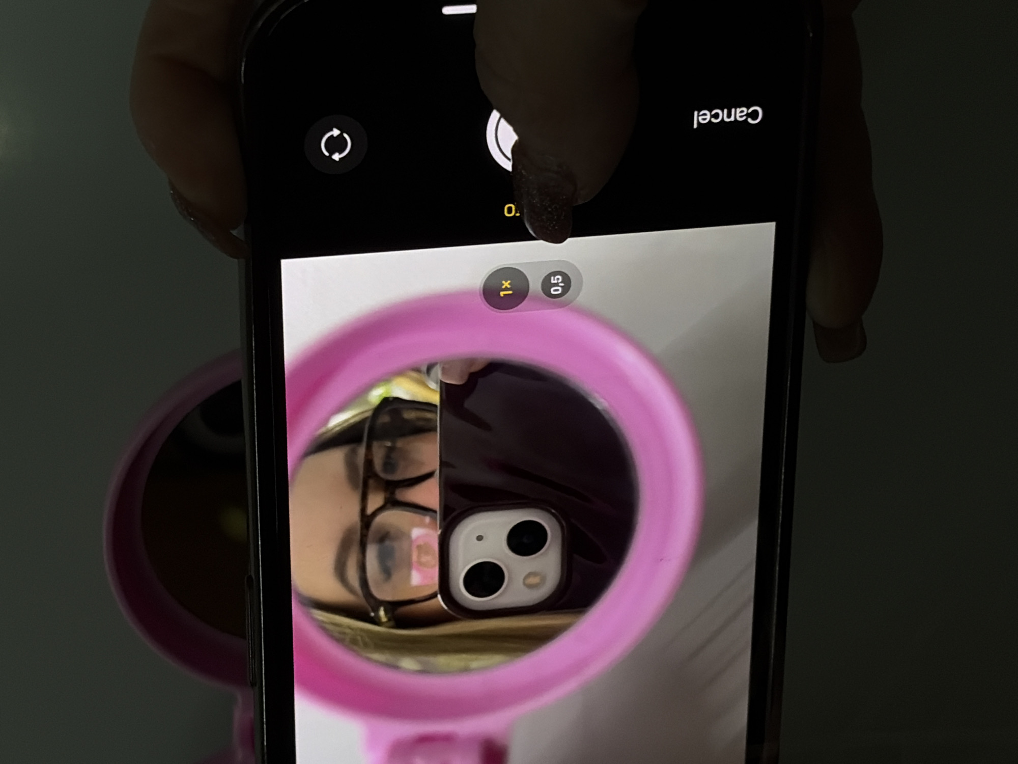 A close-up shot captures a person's hand holding a smartphone, actively displaying its camera interface, which in turn shows a reflection in a bright pink, circular mirror. The hand has polished fingernails with dark, possibly glittery, polish.
The smartphone's screen presents typical camera controls: a large white circular shutter button in the center, a circular icon with two arrows (likely for switching cameras) on the top left, and upside-down "Cancel" text on the top right. Near the bottom center, zoom options "1x" (currently selected) and "0,5" are visible.
The reflection in the pink mirror reveals a person wearing dark-rimmed glasses and with light hair. Also prominent in the reflection is the rear of another smartphone, presumably the one being held, encased in a dark purple or black cover, clearly showing a dual-camera setup with two large lenses and a flash. The design of the phone and its camera module suggests an iPhone. The scene is indoors, with the lighting primarily illuminating the phone screen and the reflection, leaving other parts of the frame in relative darkness.